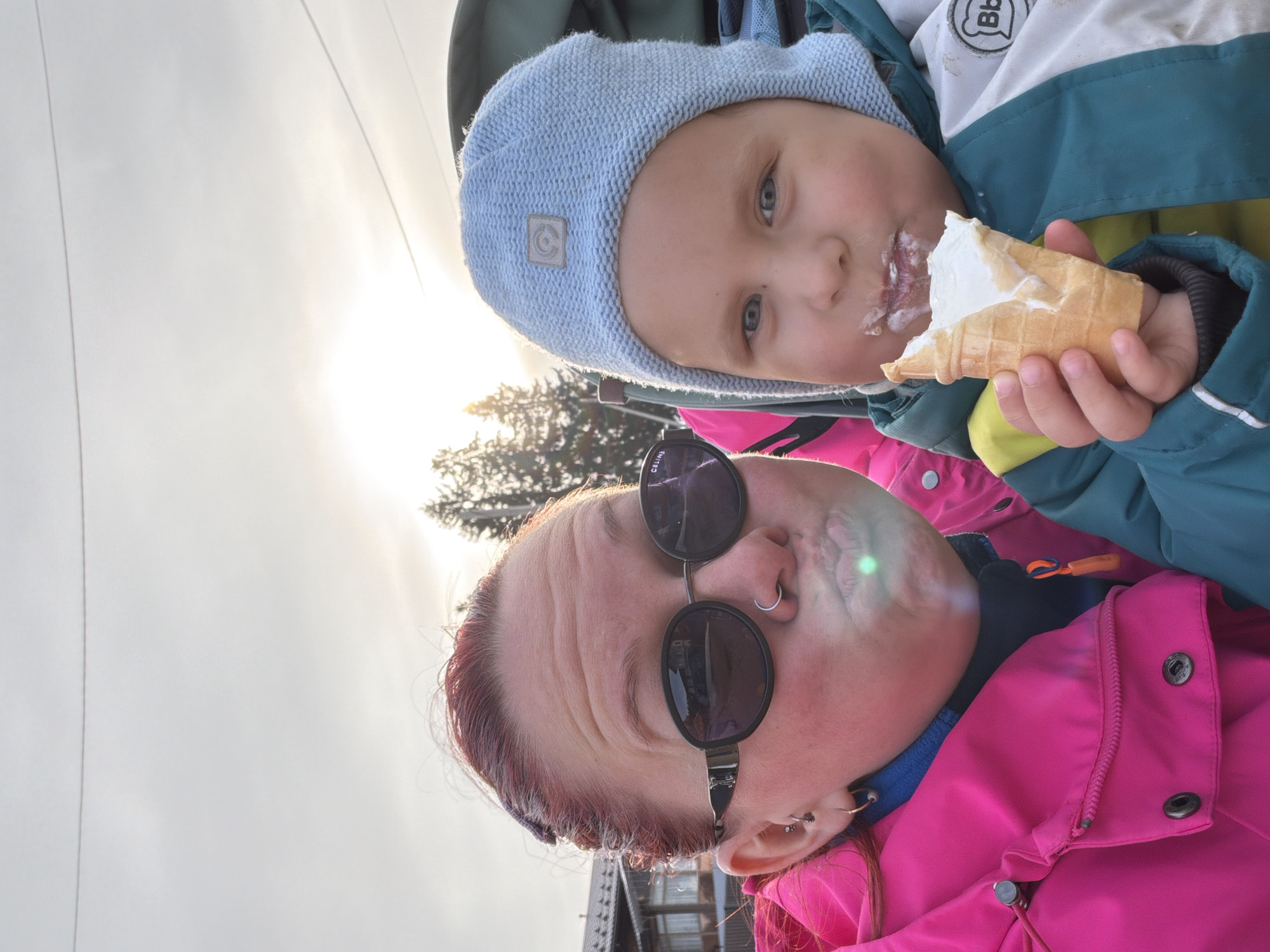 A photo shows a woman and a child. The child is eating an ice cream cone. The child is wearing a blue hat and jacket. The woman is wearing sunglasses and a pink jacket. The photo was taken in the city of Chaikovsky, Russia. A bridge structure is visible at the bottom.