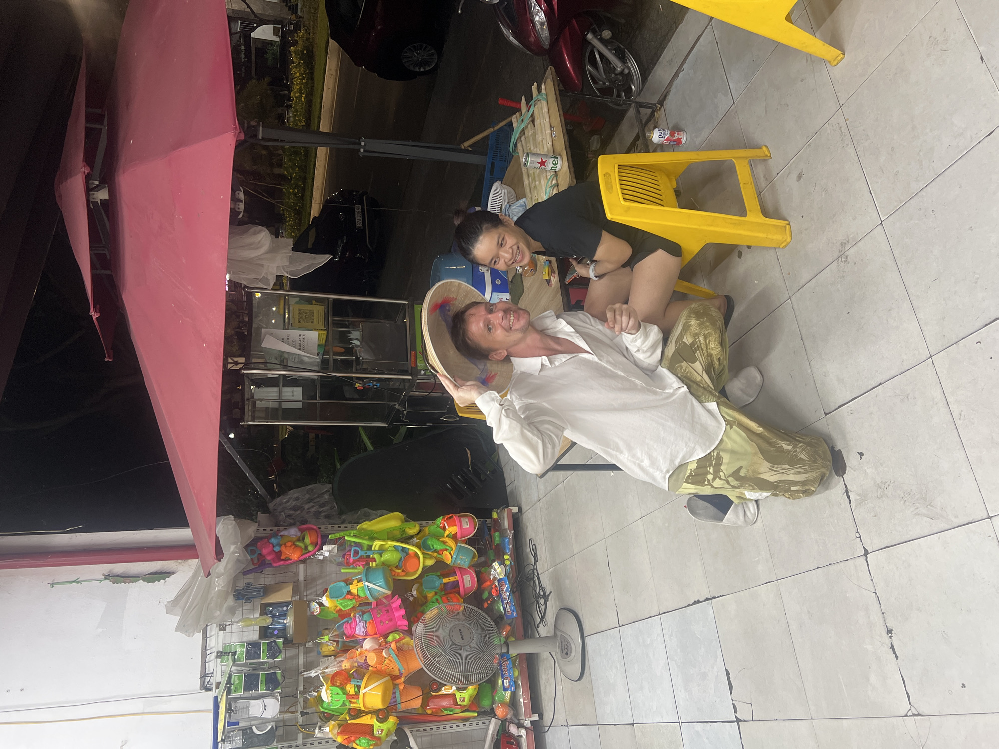 The image shows an outdoor scene at night, likely in the city of Đà Nẵng, Vietnam, where a man wearing a conical hat and a white shirt, and a woman in a black shirt are sitting at a table. The man is kneeling and posing with the hat, while the woman is seated and smiling. Behind them, a red umbrella provides shade, and shelves stocked with colorful toys and other items are visible. A standing fan is positioned to the left of the frame. In the background, there are vehicles on a street, including a red scooter. The ground is tiled, and the overall atmosphere appears to be casual and lively.