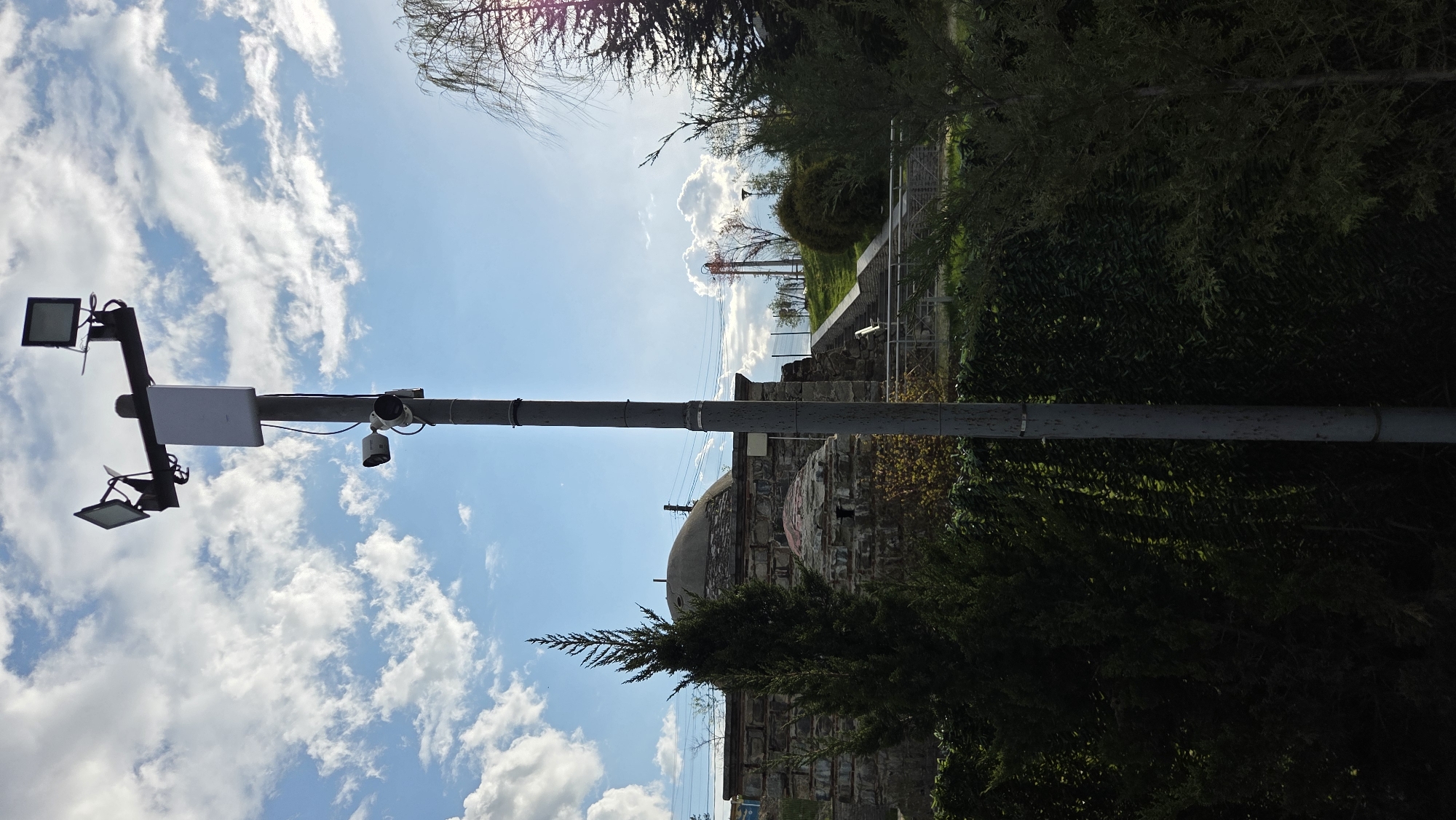 The image, oriented correctly, depicts an outdoor scene under a bright, sunny sky with scattered white cumulus clouds. A tall, grey utility pole dominates the left side of the frame, extending vertically through the middle of the composition.
Mounted on a horizontal arm extending from the utility pole is a variety of equipment. On the left side of the arm, two square, dark-colored LED floodlights are attached, one above the other, pointing downwards. Centrally located on the arm is a rectangular, flat white box, possibly an enclosure for network or electrical components. To the right of this box, two security cameras are visible: a dome-shaped camera positioned above a smaller, bullet-shaped camera, both facing outwards from the pole. Wires are visible connecting these devices along the pole.
The right side of the image features a landscape with dense green foliage, including bushes and trees. In the midground, partially obscured by an evergreen shrub, stands an old stone structure or building. It appears constructed from rough, dark-colored stones and features a weathered, grey-domed roof. Beyond this structure, a series of stone steps or a pathway leads uphill, bordered by more greenery and what seems to be a light-colored railing. In the distant background, thin power lines or communication cables are faintly visible against the bright sky.
The scene conveys a peaceful atmosphere, combining modern technological infrastructure with ancient architecture and natural elements. The time of day is bright daylight, suggesting late morning or early afternoon. There are no people visible, and no discernible text.