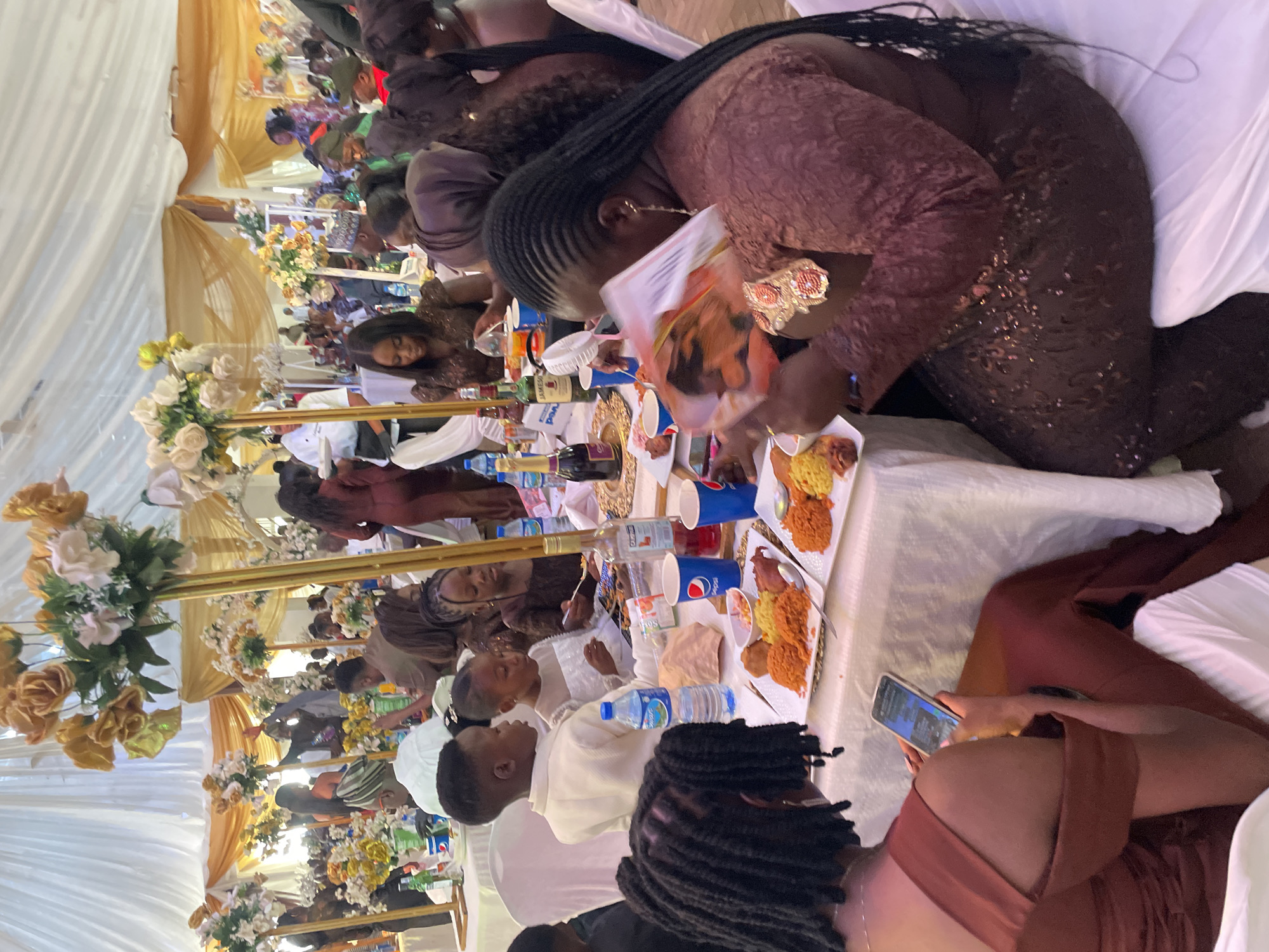 The image shows a gathering, possibly a wedding reception, taking place under a white tent structure. Tables are set with food and drinks. Individuals are seated around the tables, eating, talking, and using their cellphones. Some have braids. The tables have plates of food that may be Jollof Rice, cups, and bottles of water and alcohol. Floral decorations are visible. The location could be Jos, Nigeria.