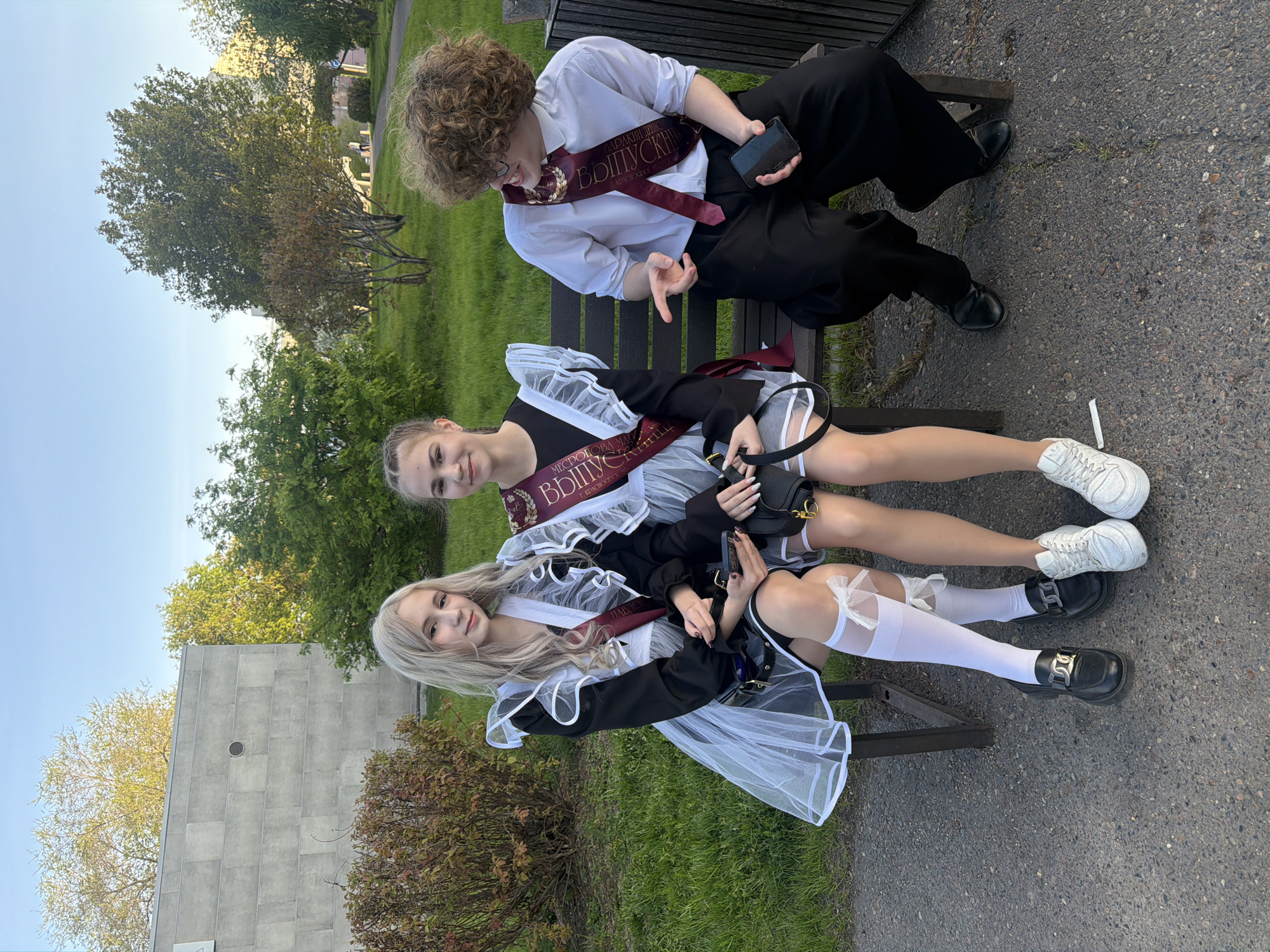 Three people are sitting on a wooden bench outdoors. Two young women are wearing black and white school uniforms with white tulle accents and red sashes that say “graduation” in Russian, while one person is wearing a white dress shirt, black pants, and a similar red sash. The young women are wearing white knee-high socks and black shoes. One is holding a black handbag. The person on the bench is holding a cell phone. The bench is situated on a paved surface next to a grassy area. Trees are visible in the background, and a stone building is also visible. The image location is in Krasnoyarsk, Russia, as indicated by the text on the sashes.