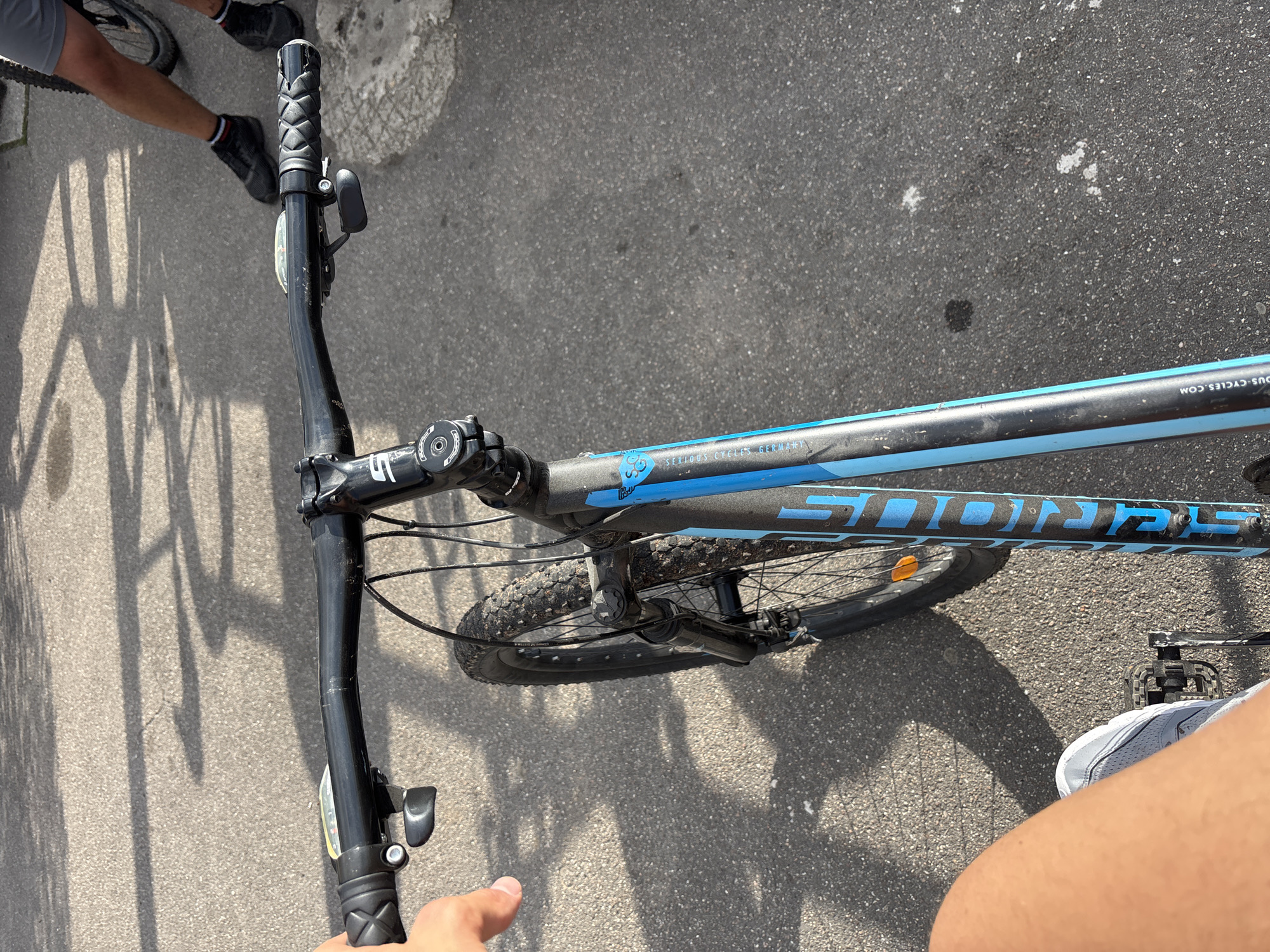 The image shows a close-up, first-person perspective of someone on a bicycle, likely in Weinheim, Germany, given the presence of "SERIOUS CYCLES GERMANY" printed on the bike frame. The focus is on the handlebars and the front wheel, with the asphalt ground visible below. Sunlight casts shadows of trees onto the road surface. A person's hand is visible on the left handlebar, and their legs and feet, wearing light-colored sneakers, are positioned near the pedals. Another person's legs and feet, wearing dark shoes and gray shorts, are visible in the background on the right side of the image, suggesting a shared outdoor space. The bicycle is black with blue accents and appears to be a mountain bike, indicated by the knobby tires and front suspension fork. The overall scene suggests a casual outdoor activity, possibly a bike ride through the city or a park.