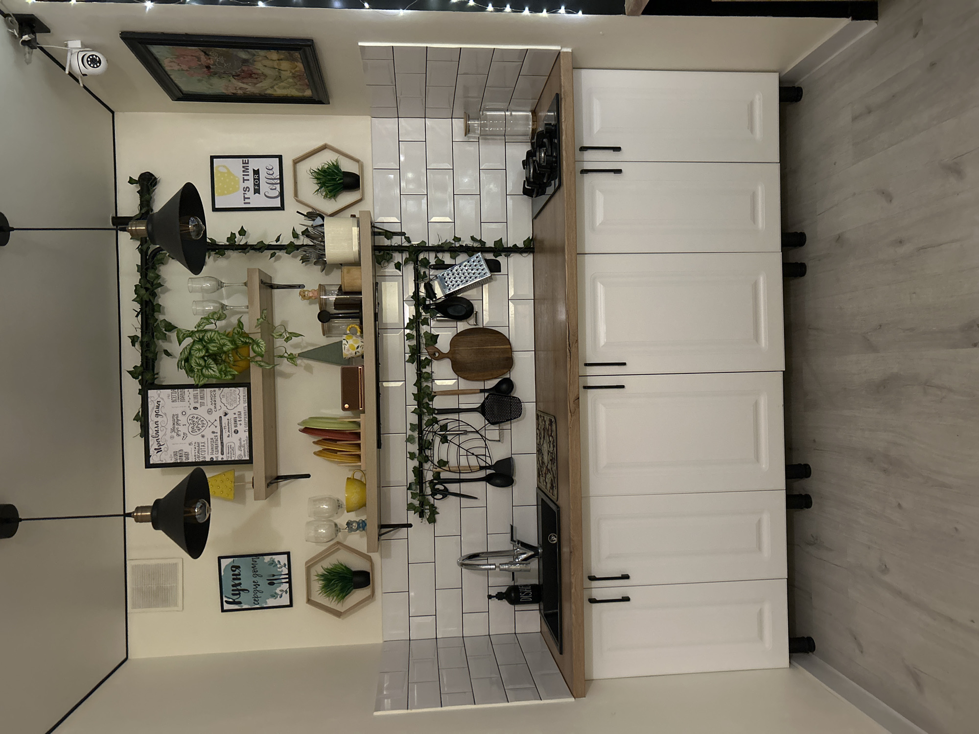 This is a photograph of a kitchen. The kitchen features white subway tile backsplashes and white cabinets with black hardware. A wooden countertop runs across the cabinets. Above the countertop are two shelves adorned with greenery. The left shelf holds a framed picture, two wine glasses, a yellow cup, and various kitchen utensils. The right shelf displays a framed picture and a hexagonal shelf with a small plant. A black sink with a silver faucet is situated on the left side of the countertop, next to a dish soap dispenser. On the right side, a gas stovetop is visible. The floor is a light-colored wood laminate. The room is illuminated by two pendant lights hanging from the ceiling. There are also some decorative elements on the wall, including framed pictures and a string of fairy lights. The scene appears to be a residential kitchen, likely in an apartment or house. There is no visible indication of the city of Bălți, Moldova, or any specific location information. The image is a still, static scene with no people or actions depicted.