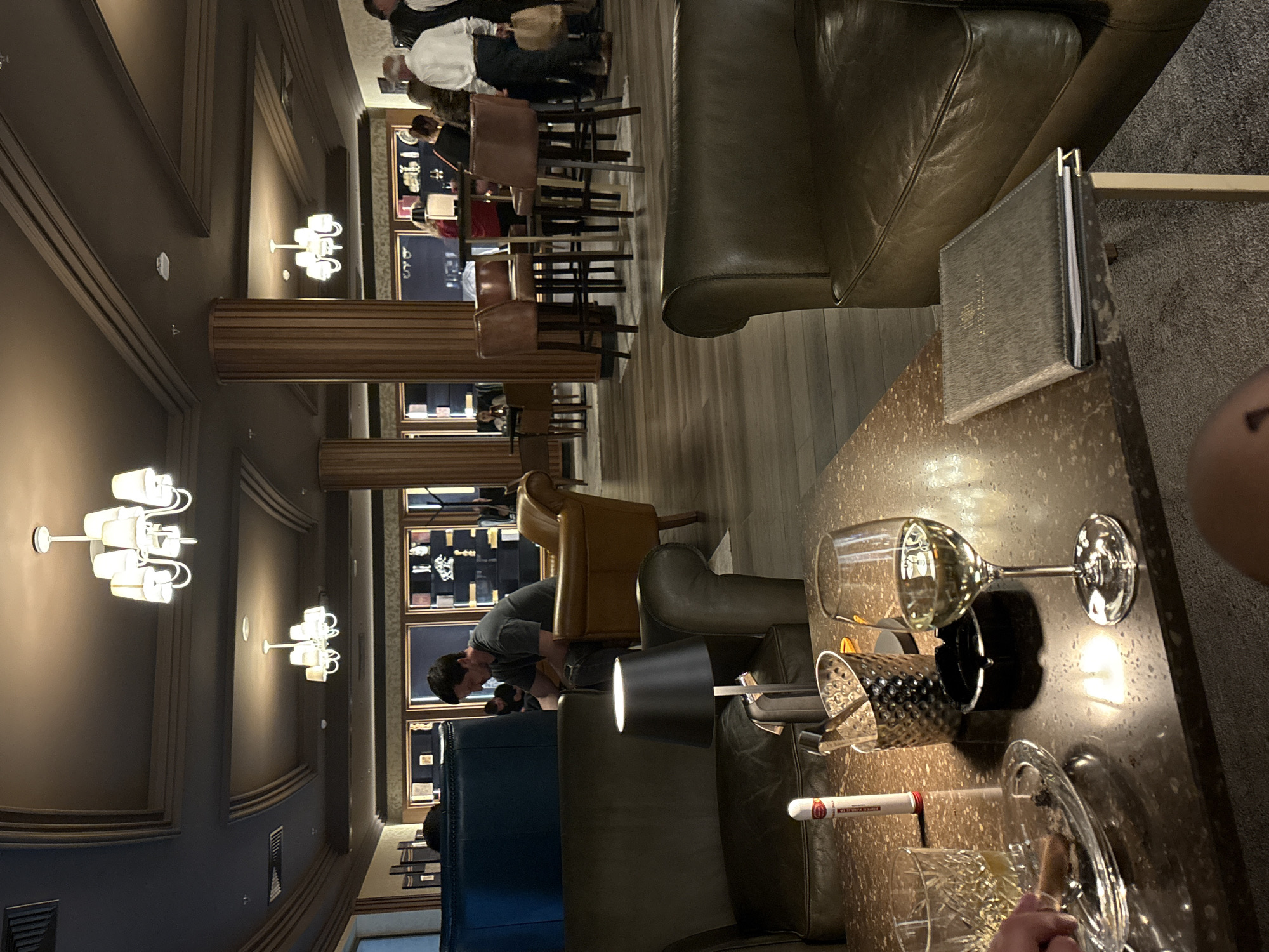 The image shows the interior of a lounge, possibly a cigar lounge. The scene includes leather seating, a bar area with high stools, and displays of items on the walls. There are people present in the bar area. A table in the foreground holds a glass of wine, a cigar, a cigar holder, and a menu or book. A lamp sits near the table. The location may be in Kazanlak, Bulgaria, but there isn't specific evidence in the image to confirm the city.
