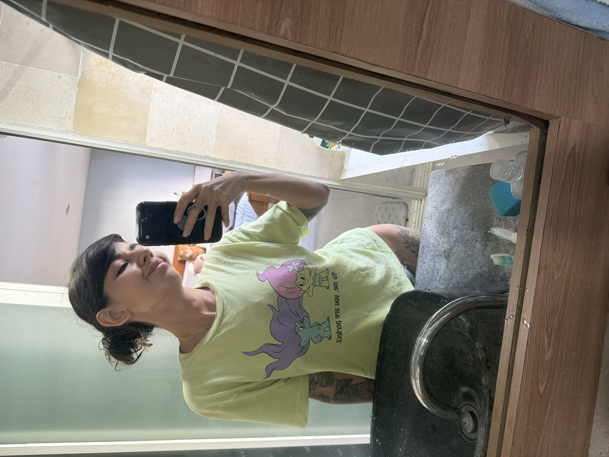 A young woman with dark hair tied up in a bun, bangs, and fair skin is taking a mirror selfie in a bathroom. She is wearing a light green t-shirt with a cartoon troll graphic and text that appears to be in reverse. She has tattoos on her left arm and thigh. The bathroom has a dark stone sink with a chrome faucet. A grey checkered shower curtain is visible in the mirror, along with a tiled wall and a wooden frame around the mirror. Several objects are on the counter, including toothbrushes, a blue sponge, and a bottle of liquid. The overall scene suggests a casual, personal moment in a home bathroom. The location is Kuta Selatan, Indonesia.