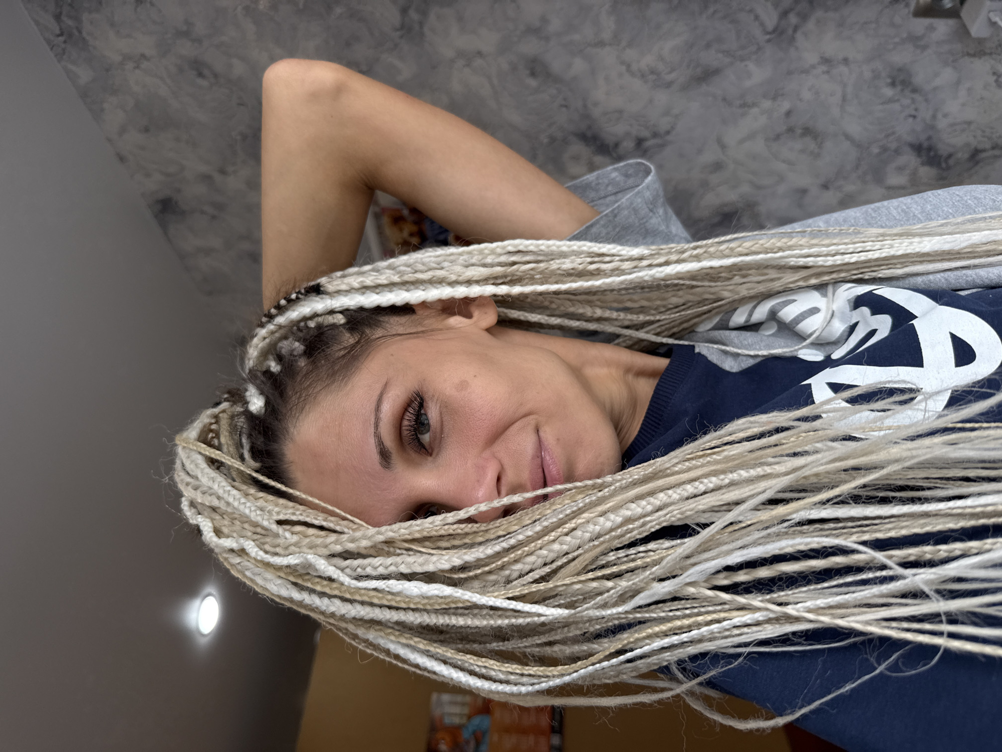 A medium shot shows a woman with blonde box braids looking to her left. Her right arm is raised, with her hand resting behind her head. She is wearing a dark blue sweatshirt. The background is a wall with a gray marbled texture. The image was taken in Rusanovo, Russia.