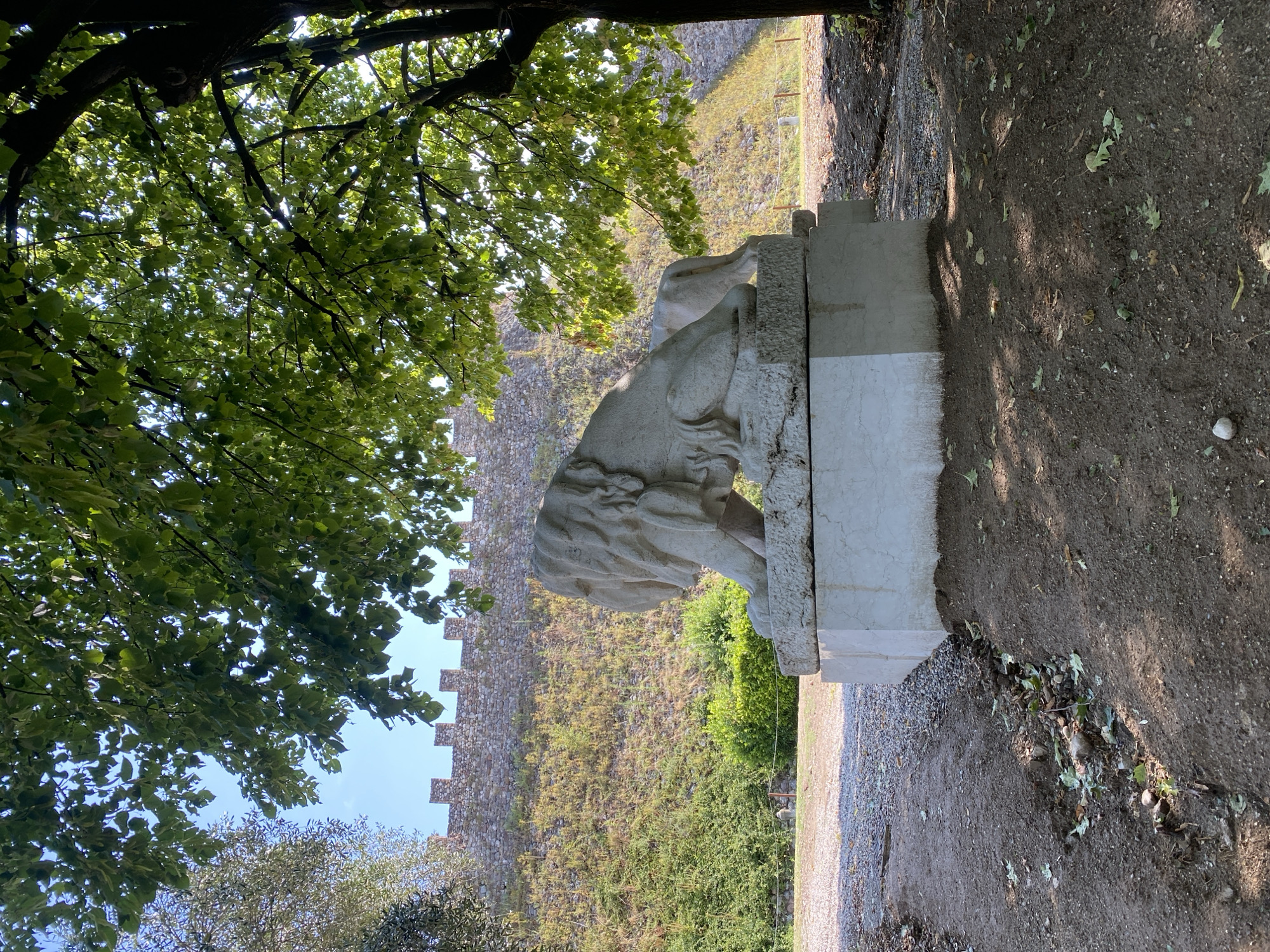A stone statue of a lion lies on a marble pedestal in the foreground, under the dappled shade of a large tree. Behind the statue, a steep, grassy embankment rises to a stone wall with crenellated battlements. The wall appears to be part of a historic fortification. The ground in front of the statue is a mixture of gravel and dark earth, with fallen leaves scattered about. The sky is clear and blue. This scene is located at the Rocca Viscontea di Lonato in Lonato del Garda, Italy.
