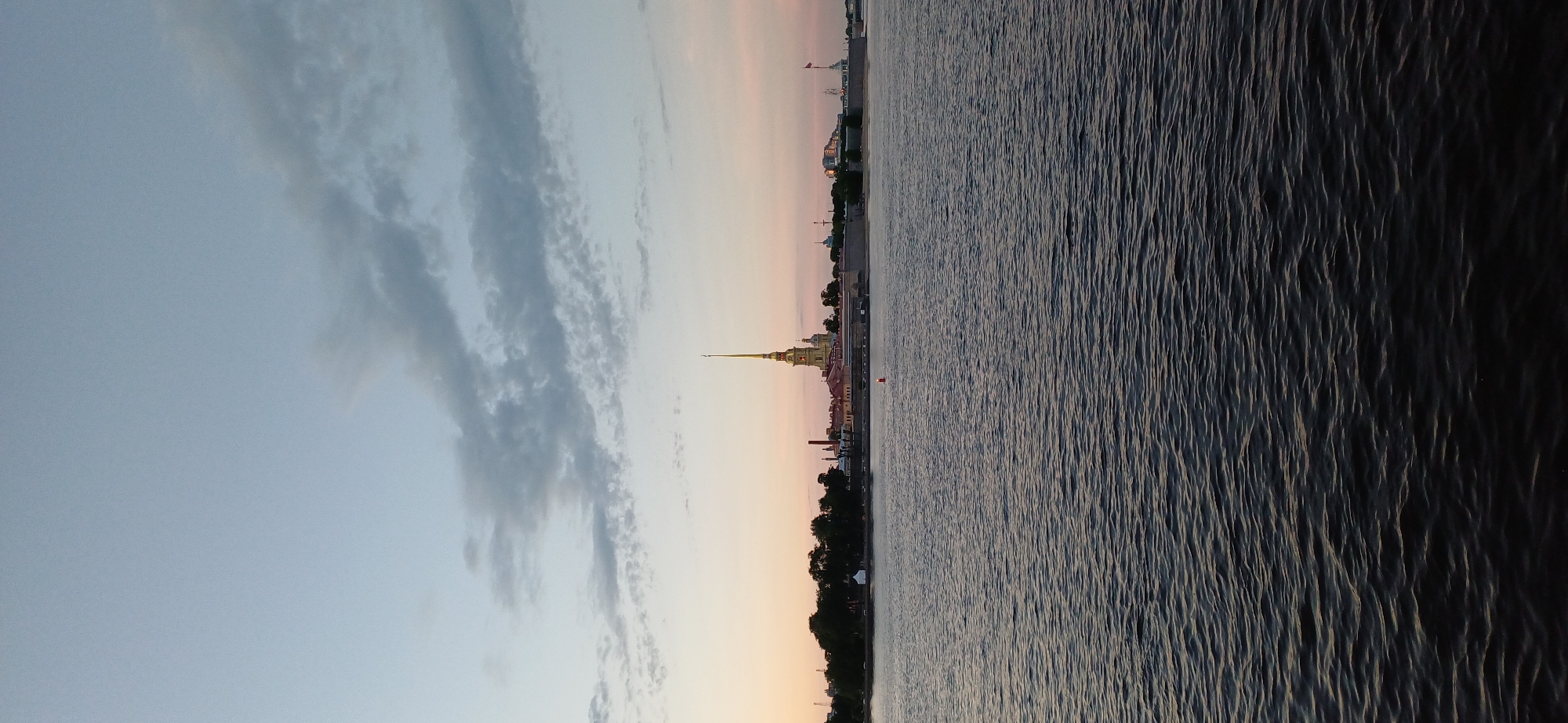 The image shows a wide body of dark, rippling water in the foreground, likely a river or gulf. In the middle ground, a long, low embankment or wall lines the water's edge, with a line of trees behind it. Dominating the skyline above the embankment is a prominent yellow building with a tall, slender spire topped with a golden angel. This is characteristic of the Peter and Paul Fortress, a historical landmark in Saint Petersburg, Russia. Several other buildings, some with red rooftops, are visible along the embankment. The sky above is a gradient of soft colors, transitioning from pale blue at the top to a light pink and orange near the horizon, suggesting either sunrise or sunset. A few wispy clouds are scattered across the sky. The scene is serene and appears to capture a moment of twilight. There are no people or discernible actions depicted in the image.