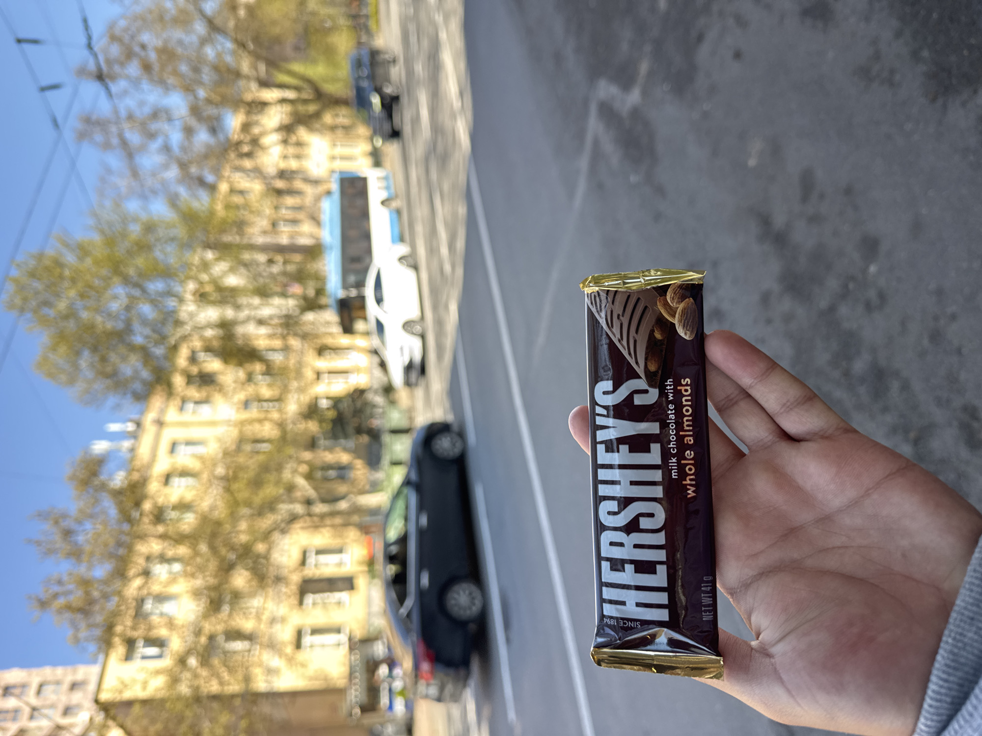 A person holds a Hershey's milk chocolate bar with whole almonds. In the background, there is an urban scene. A tall building with yellow walls and multiple windows is on the left, partially obscured by bare trees. Cars are parked along the street, and a bus is also visible. The picture was taken in Yerevan, Armenia.