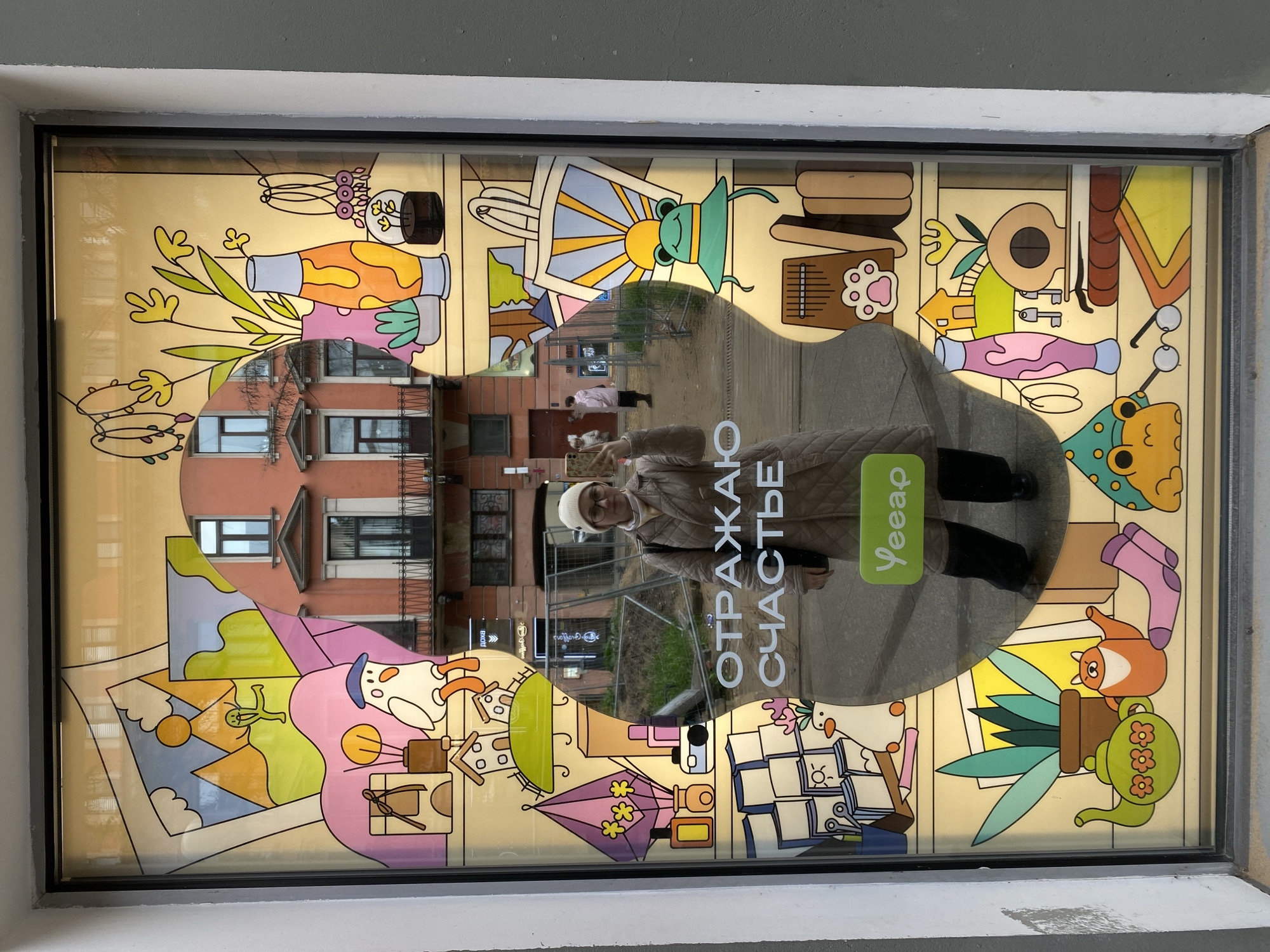 Here is a description of the image:
The image captures a storefront window in Saint Petersburg, Russia. The window is decorated with a colorful, cartoonish mural featuring various whimsical objects and characters. The mural is predominantly yellow, with details in pink, orange, green, and blue.
A circular section of the window is clear, providing a view of the street outside. Reflected in this clear area is a person holding a phone, presumably taking the picture. The person is wearing a beige jacket and a white hat. The street scene includes a building, a fenced-off area, and another person walking in the distance. Text in Russian is visible near the reflected person. A logo, seemingly for a delivery service, is also visible.