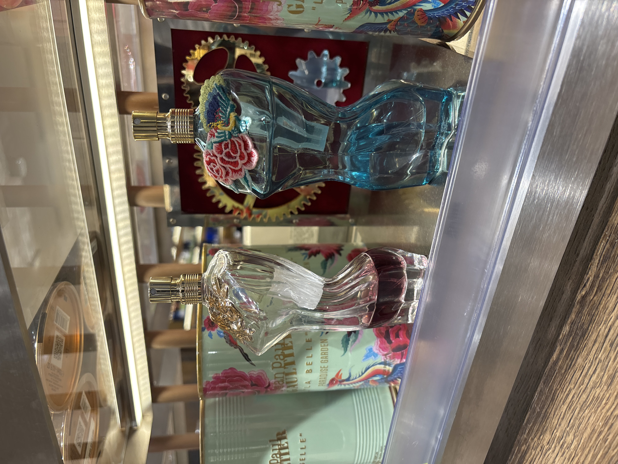 The image shows a display of perfume bottles, likely in a retail setting. Two distinct perfume bottles are visible: one with a clear bottle and gold accents, and another with a blue bottle and embroidered floral details. The bottles are displayed on shelves, with the product packaging visible behind them, including the brand name "Jean Paul Gaultier" and the words "La Belle Paradise Garden." The background includes metal gears and other decorative elements. The overall setting appears to be a well-lit, organized retail space. I cannot determine the location as Saint Petersburg, Russia, with the information provided.