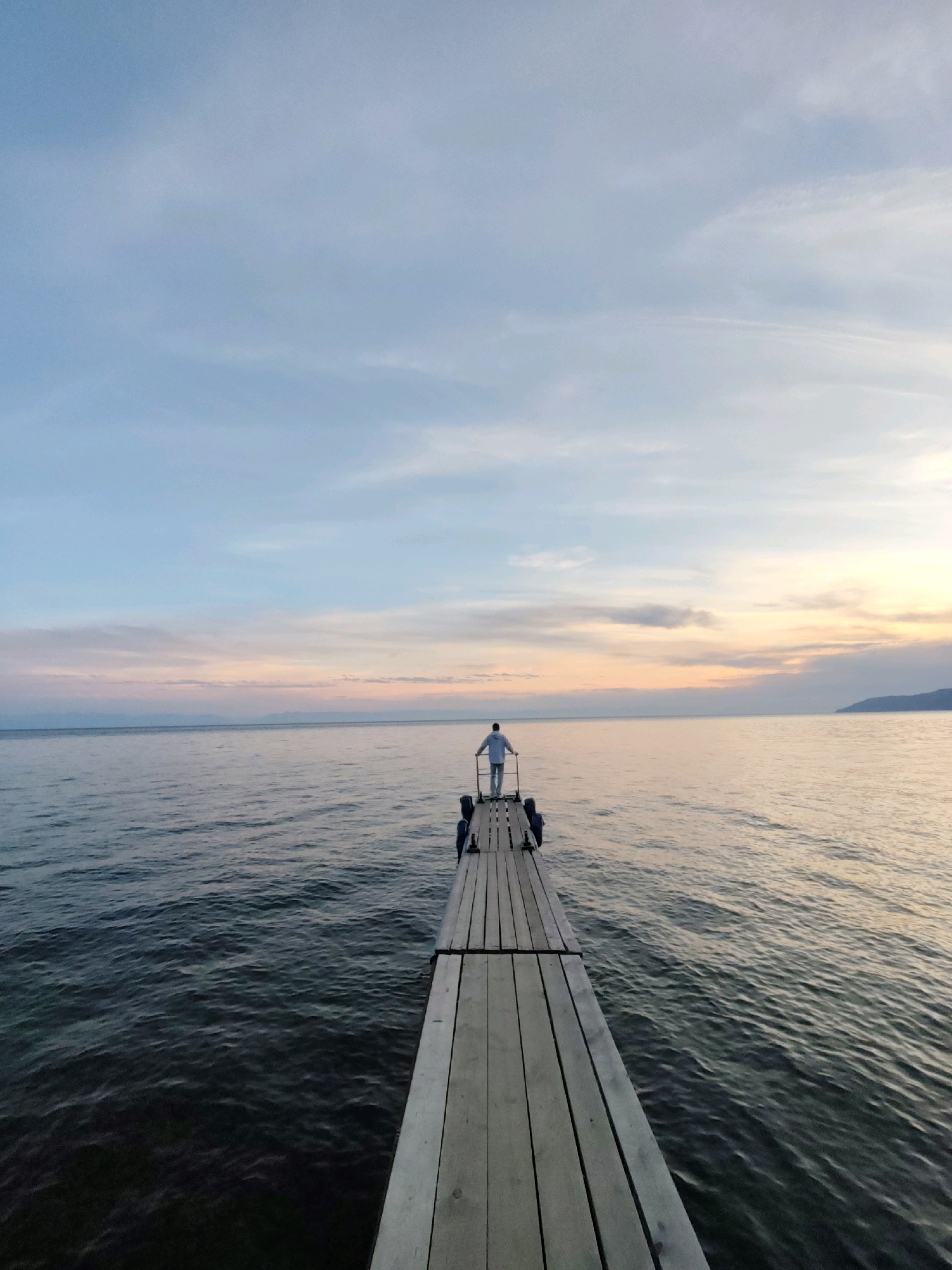 An individual stands on a wooden pier extending into a calm body of water under a sky with soft hues of blue, pink, and orange, indicative of dawn or dusk. The pier is constructed of weathered wooden planks and has a railing on either side. The person, dressed in light-colored clothing, faces away from the camera, looking out towards the horizon. The water exhibits subtle ripples, reflecting the colors of the sky. In the distance, a faint silhouette of land or mountains can be seen along the horizon. This scene appears to be set on Lake Baikal in Listvyanka, Russia, a popular tourist destination known for its stunning natural beauty and its pier infrastructure.