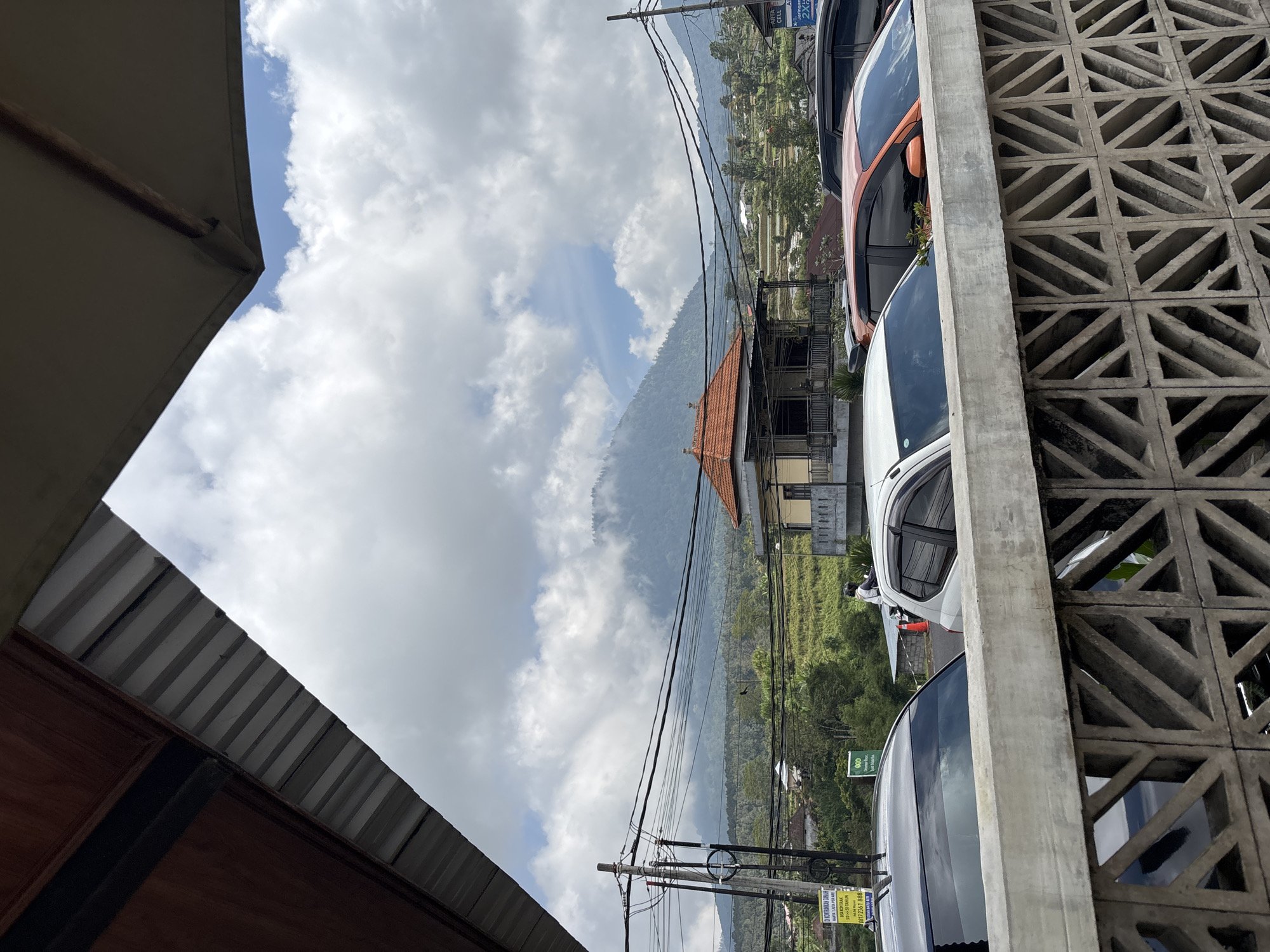 The image shows a view in Baturiti, Indonesia, with a mountainous landscape in the background partially obscured by clouds. A traditional Balinese building with a red tiled roof is situated in the middle ground. Foreground elements include vehicles parked alongside a patterned concrete wall on the right, and a partial view of a roof or awning on the left. Power lines stretch across the middle of the frame. The overall perspective is tilted or rotated.