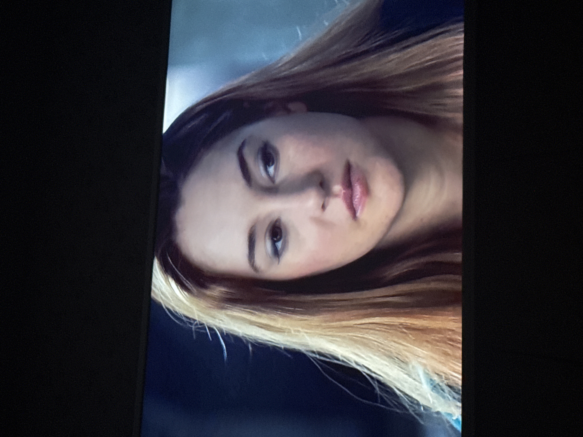 The image is a close-up shot of a woman's face. She has fair skin, light brown hair with blonde highlights, and dark eyebrows. She is looking directly at the camera. The background is blurred and dark, with hints of blue and gray. The photo appears to be taken indoors. The location is not discernible from the image.
