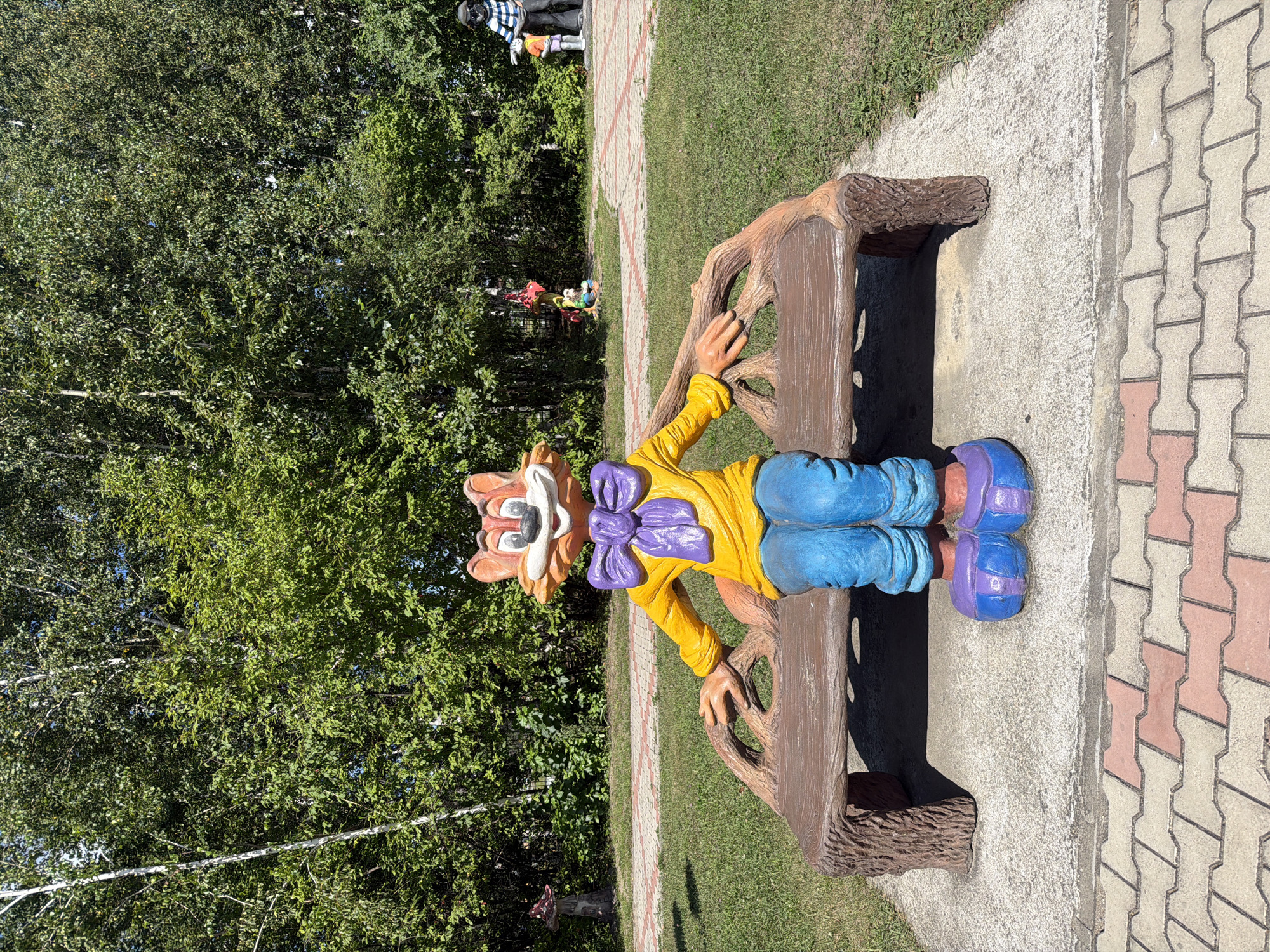 In Ivanovka, Russia, a colorful cartoon fox sculpture sits on a wooden bench in a park. The fox is wearing a yellow shirt, blue pants, and purple shoes, with a large purple bow around its neck. The bench has a rustic, wood-like design. Behind the fox, a paved path with red and tan interlocking stones leads through a grassy area surrounded by trees. In the background, other whimsical sculptures are visible, including a mushroom-shaped one to the left and figures of people and animals further back. The scene is illuminated by bright sunlight, indicating a clear day.