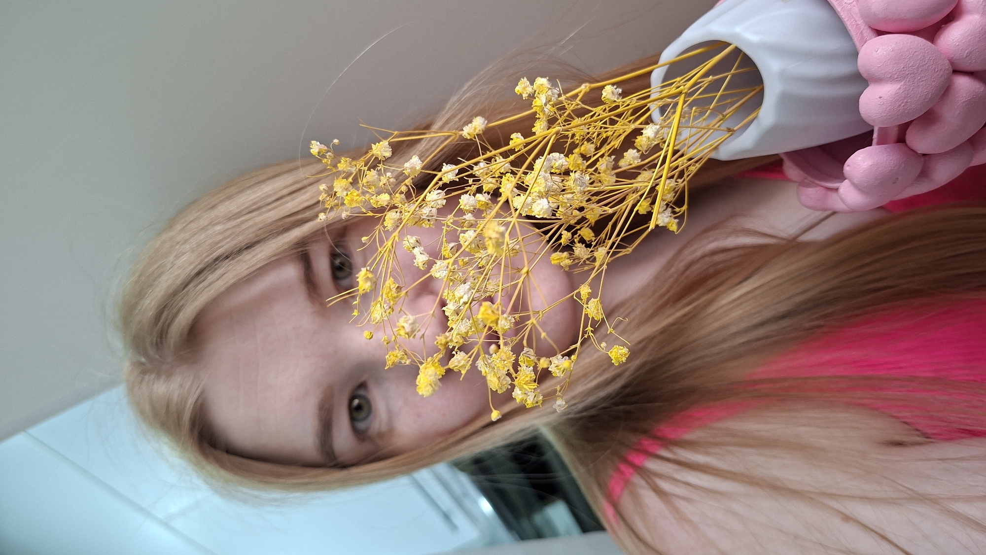 A close-up, angled shot shows a young woman with long, light brown hair partially obscuring her face with a bouquet of small yellow flowers. The woman's eyes are visible through the flowers. A light blue vase, partially covered with pink, heart-shaped objects, holds the flowers. The woman is wearing a bright pink top. The background appears to be a light blue or gray surface, potentially a wall or table. The location is specified as the city of Chaikovsky, Russia.