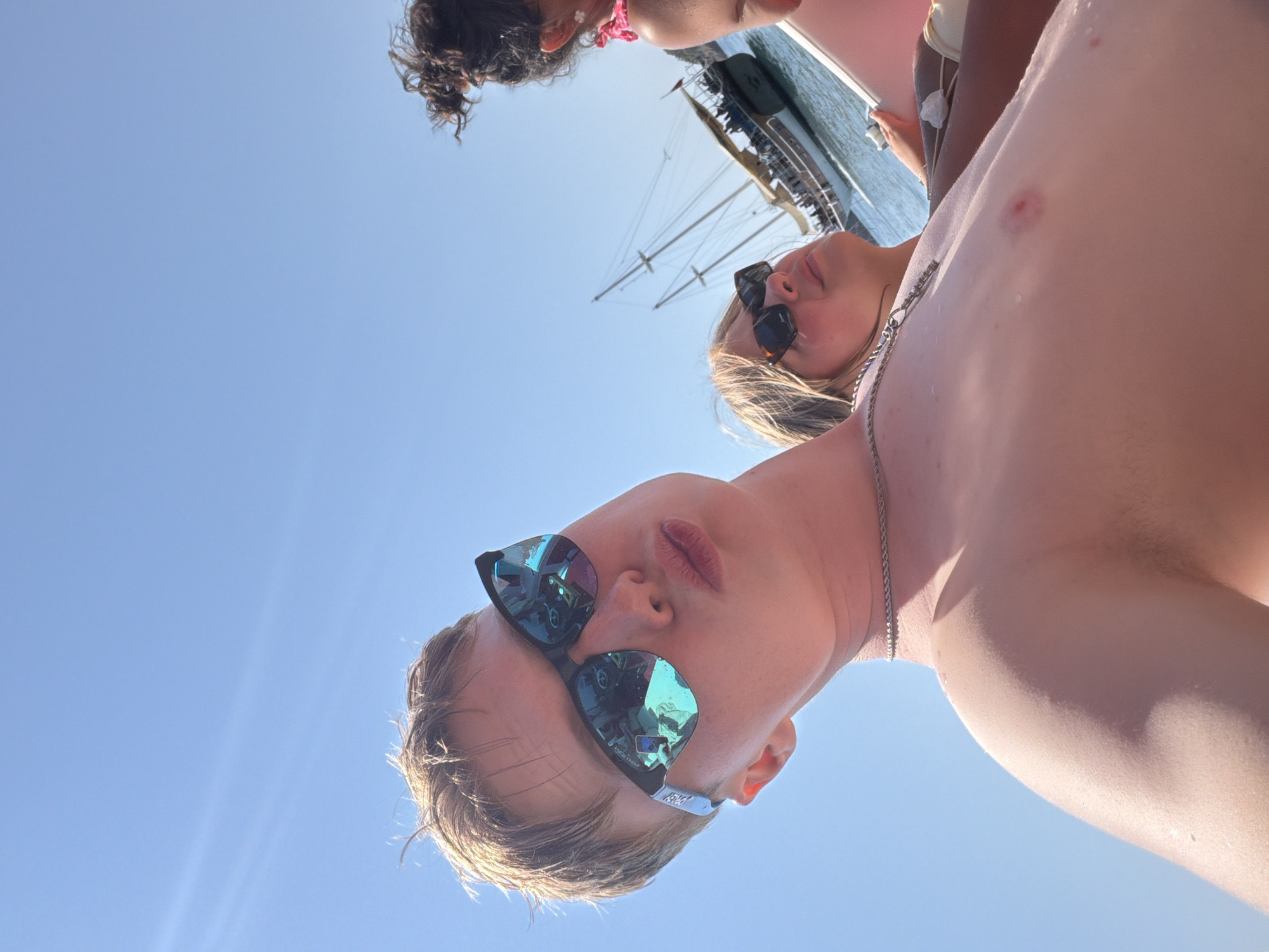 This is a close-up, low-angle, point-of-view photograph taken outdoors on a sunny day, likely in or near Kemer, Turkey. The focus is on a young person in the foreground wearing sunglasses and looking slightly upwards, with their lips pursed. Their chest is visible, showing a light chain necklace. To the left of the frame, another young person is visible wearing sunglasses and looking upwards. In the background, a large sailing boat with multiple masts and sails is on the water, with other people visible on its deck. The sky is clear and blue. The overall impression is a casual, leisure activity, possibly a boat trip on the Mediterranean Sea.