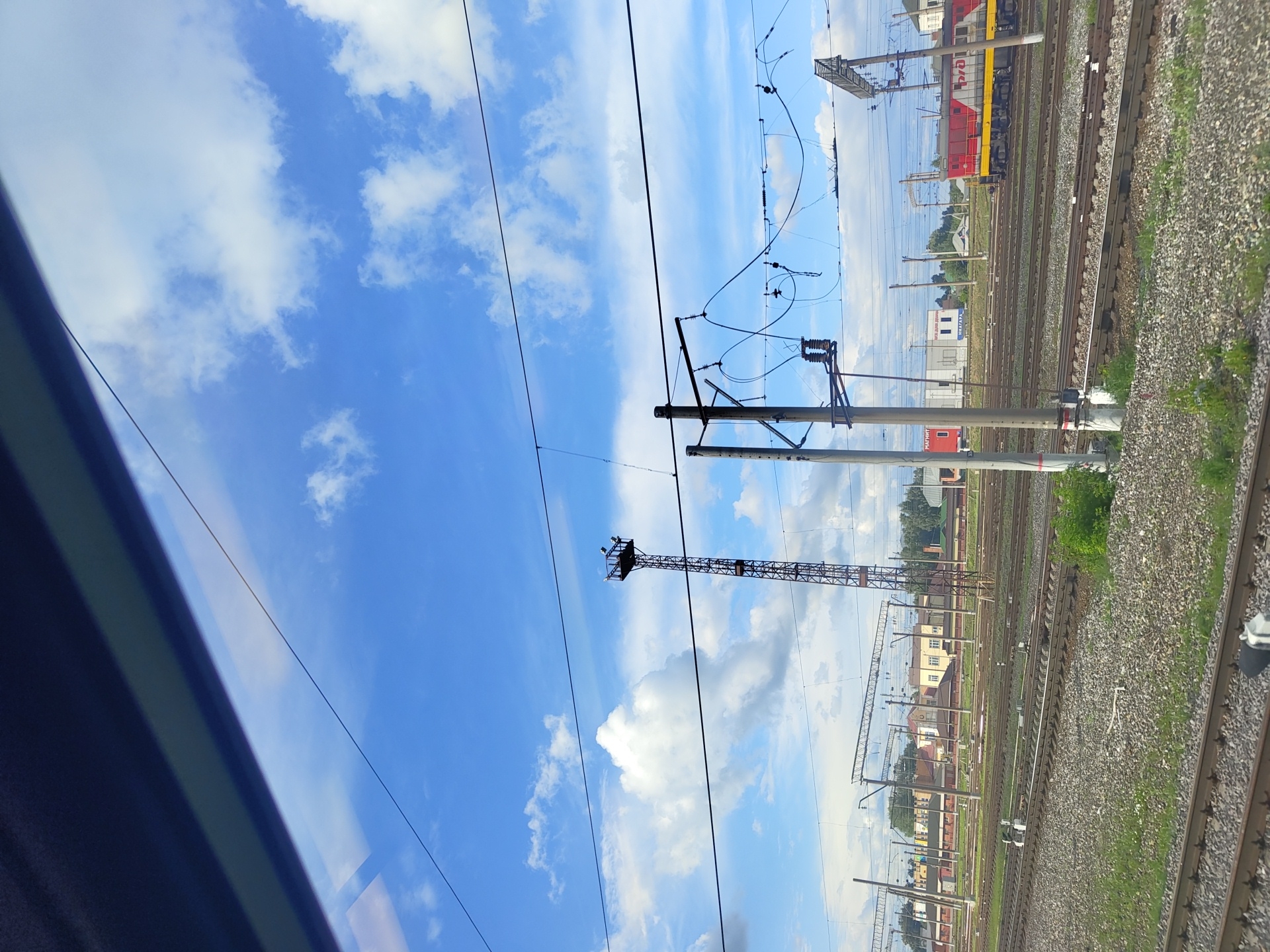 The image shows a view from a train window, looking out at a railway yard under a blue sky with scattered clouds. In the foreground, there are multiple sets of train tracks covered with gravel and some sparse vegetation. Several utility poles with overhead wires are visible throughout the yard, some with complex wiring systems and insulators. A tall, lattice-like tower with lights at the top stands prominently in the middle ground. In the background, there are various buildings, including what appear to be small industrial or service buildings with signs, and some residential-looking structures. A red and yellow train engine is parked on one of the tracks in the distance, to the right of the frame. The overall scene depicts an active railway operational area within a city, likely Фруктовая, Russia, given the context. There are no people clearly visible in the image.
