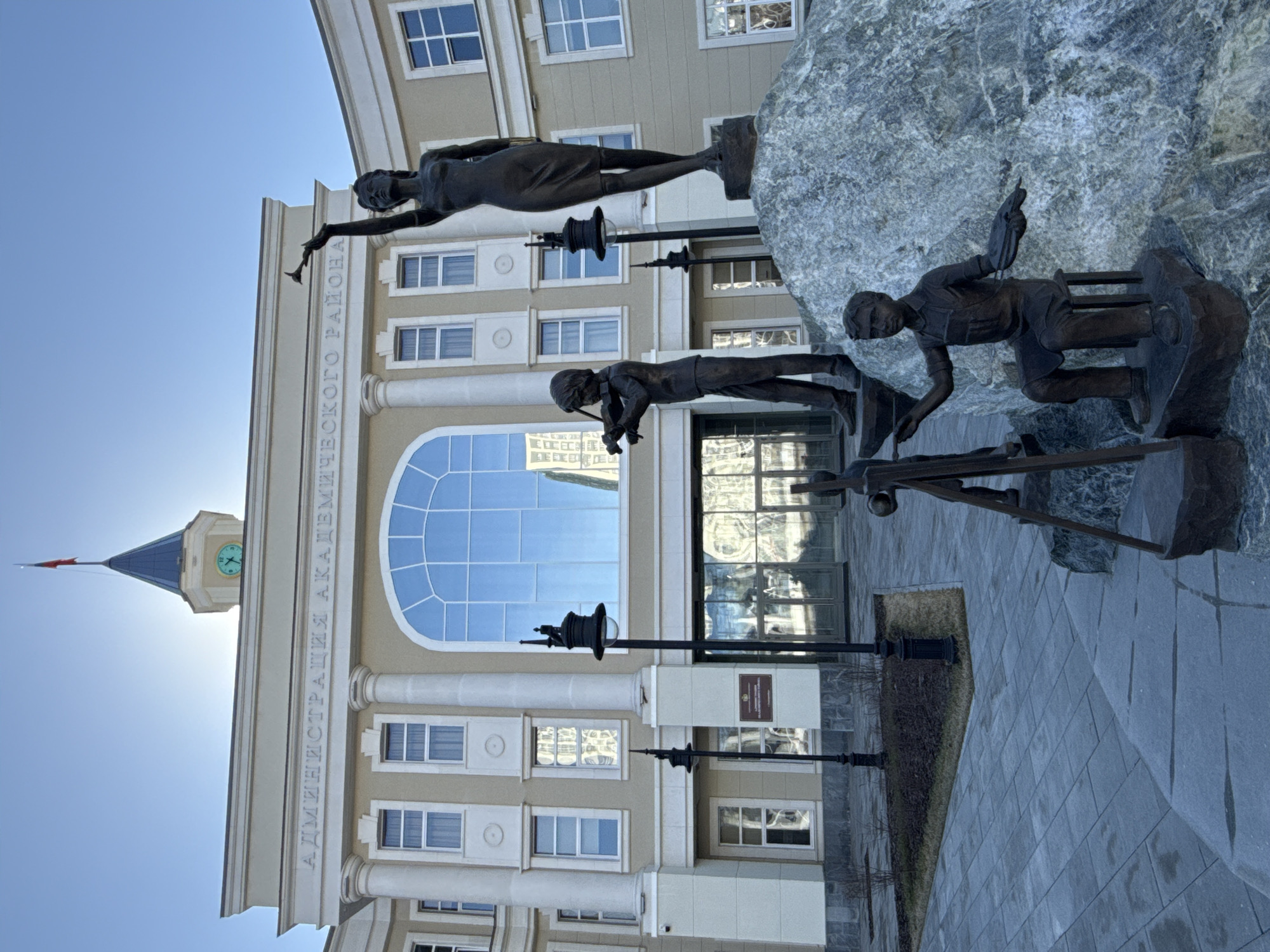 The image depicts a sculpture outside the Administration Building of the Academic District in Yekaterinburg, Russia. The sculpture consists of three bronze figures mounted on a large rock. The topmost figure is a woman standing on a pedestal, holding her arm out. The middle figure is a man, also on a pedestal, appearing to be working with tools. The bottom figure is a child sitting and working on a project. The Administration Building behind the sculpture is a multi-story, light-colored building with classical architectural elements, including columns and a large arched window. A clock tower is visible on the left. Lampposts are positioned in front of the building. The ground is paved with gray stones. The sky is clear and light blue.