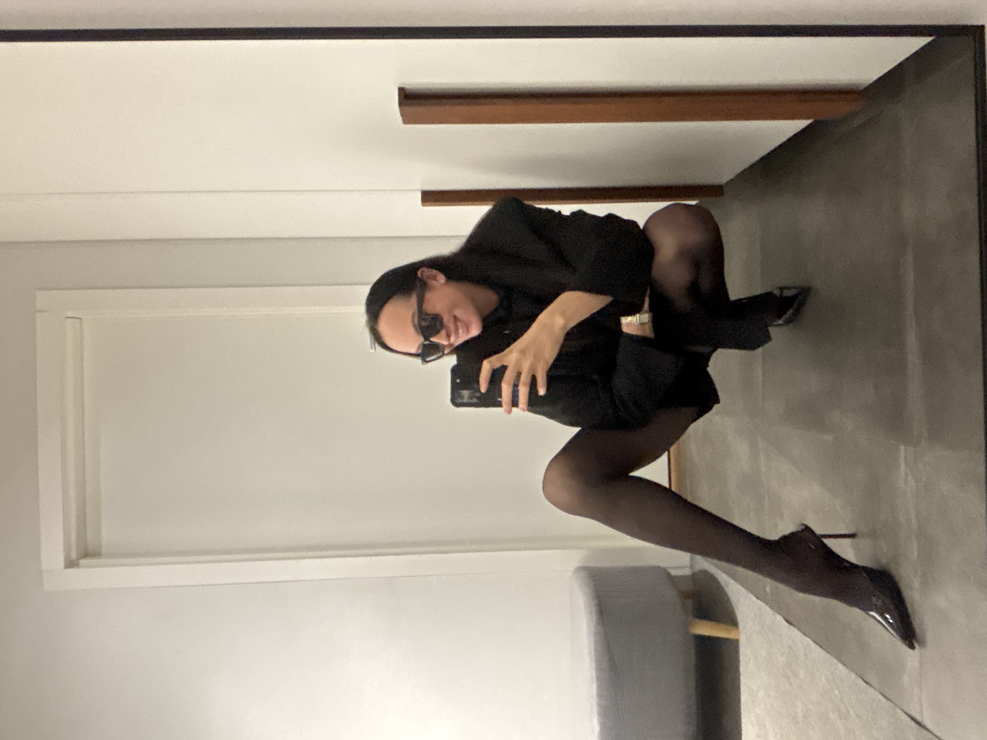 The image displays a mirror selfie of an individual in a squatting posture within an indoor setting in Chaikovsky, Russia. The subject, a person with dark hair, is wearing black square-frame sunglasses, a dark blazer-style top, black sheer tights, and black pointed-toe stiletto mule heels. A gold-toned wristwatch is visible on their left wrist. The individual is smiling and holds a dark-colored smartphone in their right hand, aimed at the mirror. The background reflection reveals a dark gray floor, light-colored walls, a closed white door with a white frame on the left, and a partially visible light gray circular ottoman with light wooden legs. Two vertical wooden panels are affixed to the wall on the right side of the frame.