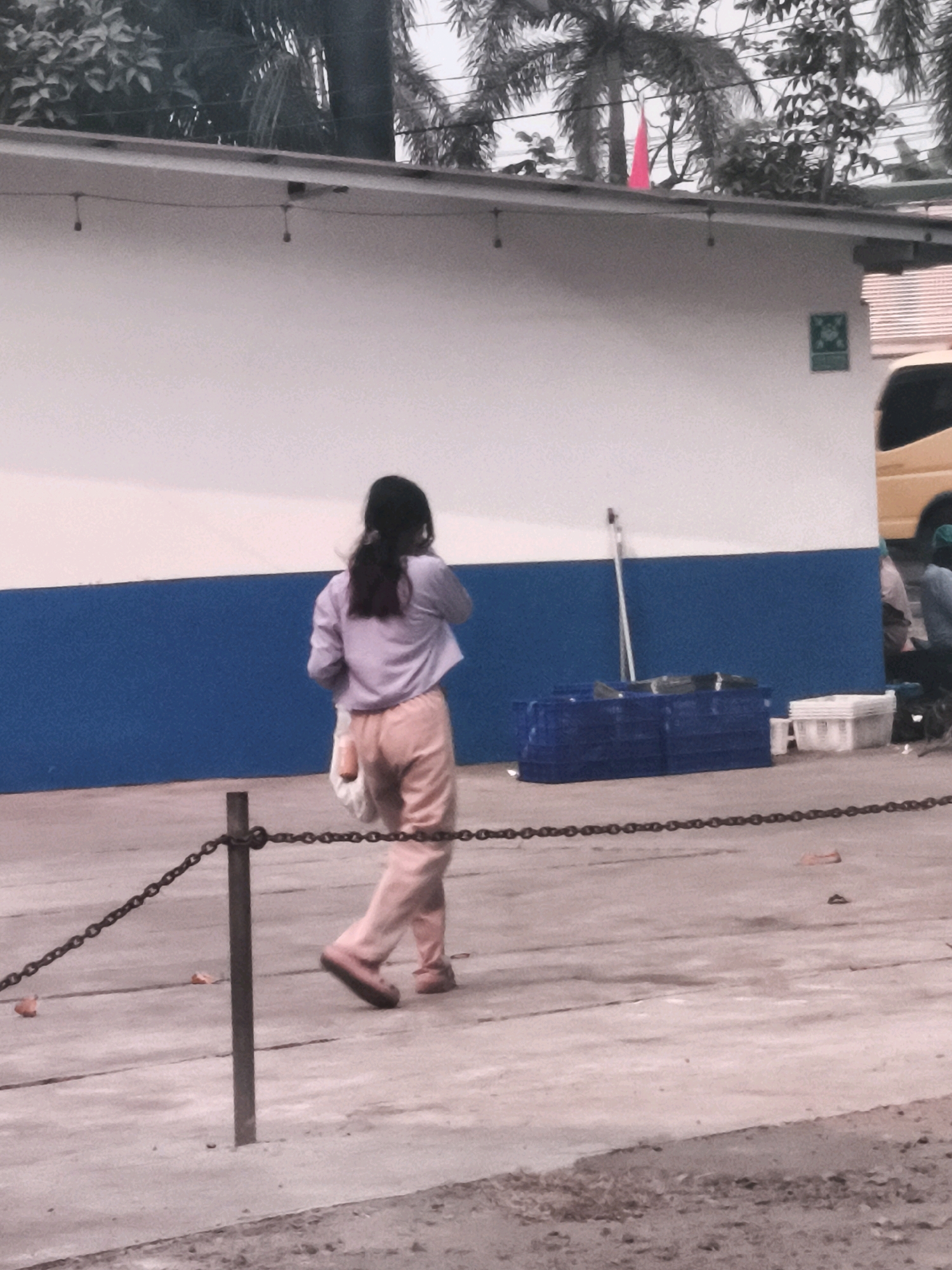 The image depicts an outdoor scene in Palembang, Indonesia. A young person, viewed from the back, is walking on a paved surface. They are wearing a light purple long-sleeved shirt and pink pants. A white plastic bag is being carried in their right hand. A chain fence with a metal post runs across the foreground. In the background, there is a building with a white upper half and a blue lower half. To the right of the building, a portion of a yellow vehicle is visible. Several blue plastic crates and white containers are stacked on the ground near the building. Trees and foliage are present above the building's roof. The lighting suggests it is daytime.