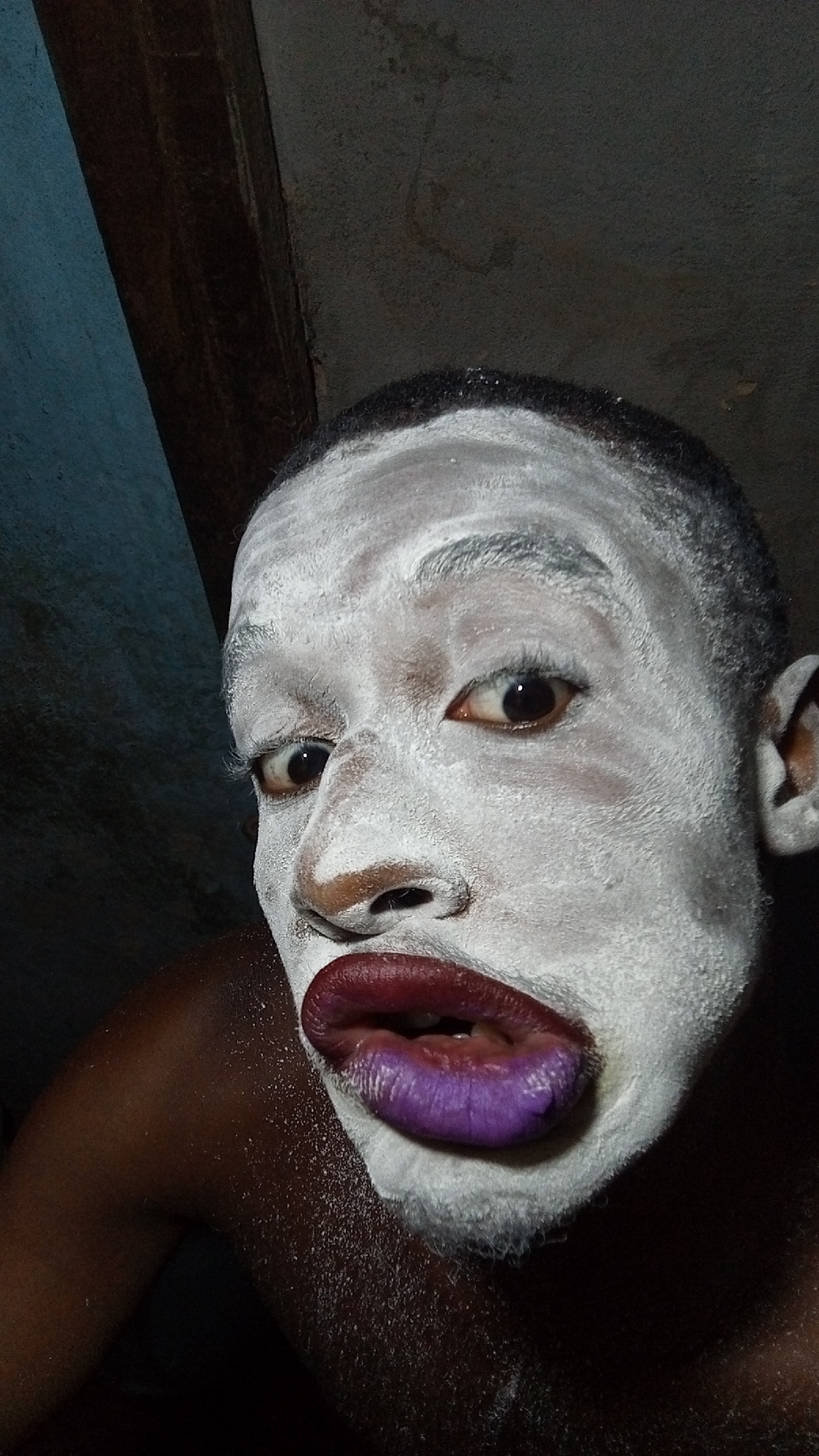 A close-up portrait features a person with dark skin, their face heavily covered in white powder or paint. Their lips are strikingly colored, with a dark reddish-purple on the upper lip and a vibrant purple on the lower lip, which is slightly parted. Their dark eyes are wide open, looking directly at the camera with an intense or theatrical expression. Short, dark hair is visible at the top of the forehead, also dusted with white powder. The person's bare, dark-skinned shoulder and upper arm are visible on the left side, showing some texture. The background is dark and indistinct, with hints of what appears to be a rustic or unadorned wall structure on the upper left, suggesting an indoor setting with low illumination. No text is visible in the image.