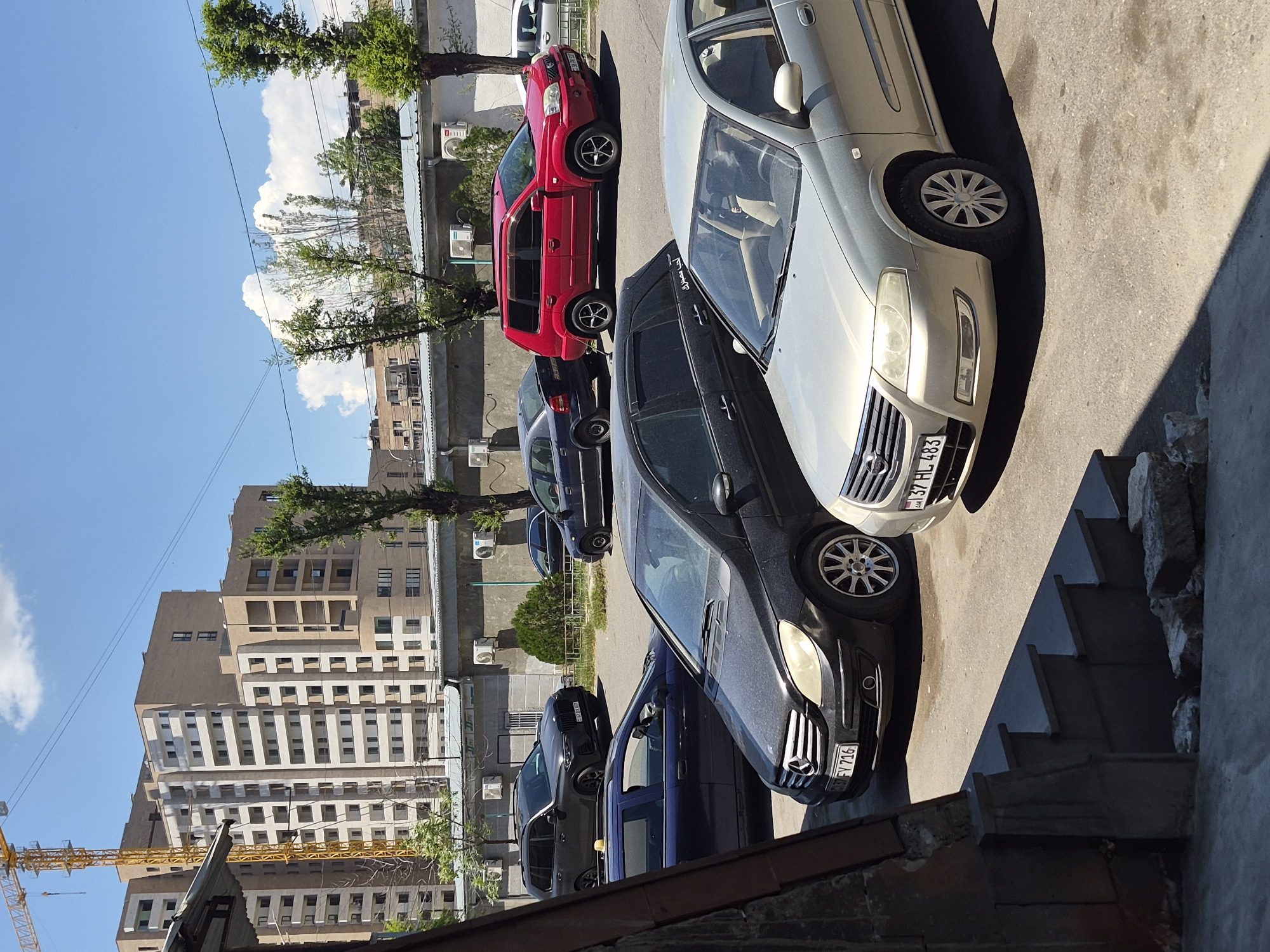 The image captures a sunny urban scene in Yerevan, Armenia, showcasing several parked cars alongside a street or parking area. The time of day appears to be mid-morning or afternoon, indicated by the strong shadows cast by vehicles and structures.
In the foreground, a silver or light gold sedan, identifiable as a Nissan (likely a Sunny or Almera) from its grille and partial brand visibility on the license plate, is parked. Its Armenian license plate reads "37 HL 483." Behind it, a dark grey or black Mercedes-Benz sedan is parked, displaying the license plate "50 FV 716." Further down the line, a blue hatchback, a red SUV (likely a Nissan X-Trail) with the license plate "58 AR 051," and another dark blue sedan are visible. The cars are parked on asphalt, which features distinct shadows.
In the background, a diverse urban landscape unfolds. A large, multi-story modern building with numerous windows stands prominently, alongside other residential and commercial structures. Green-leafed trees are scattered throughout the scene, lining the street and framing the buildings. A bright blue sky with large, fluffy white clouds dominates the upper portion of the image. To the far left, a yellow construction crane is visible against the sky, hinting at ongoing development. Air conditioning units are affixed to the facades of some buildings. The scene conveys a clear, bright day in a lively city environment.