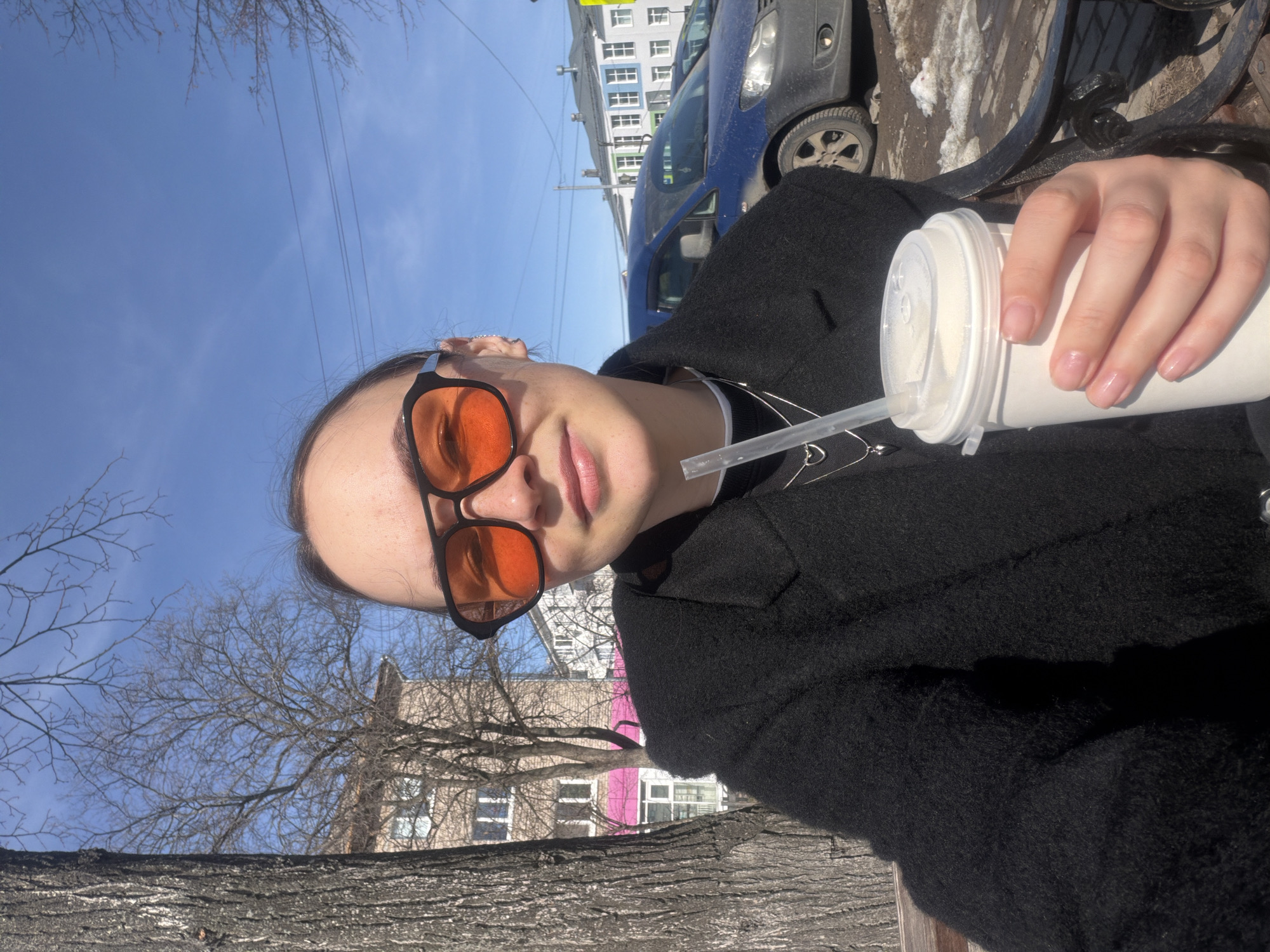 A person wearing orange-tinted sunglasses and a black coat holds a white cup with a straw. In the background, there is a blue sky with bare trees, a building with white window frames, and a blue van. The image was taken from a low angle. A bench is partially visible at the bottom of the frame. Based on the buildings and architecture, the location is in the city of Chaikovsky, Russia.
