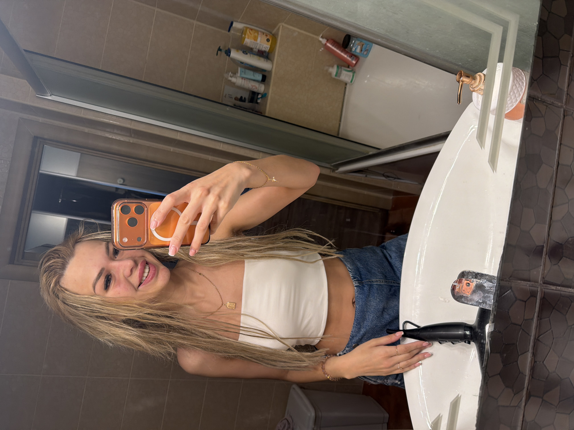 In Kyiv, Ukraine, a young woman with blonde hair, wearing a white tube top and denim shorts, takes a selfie in a bathroom. She is smiling and holding an orange smartphone in her right hand. Her left hand rests on the edge of a white sink. The bathroom features beige tiled walls, a glass shower door, and various toiletries on a shelf.