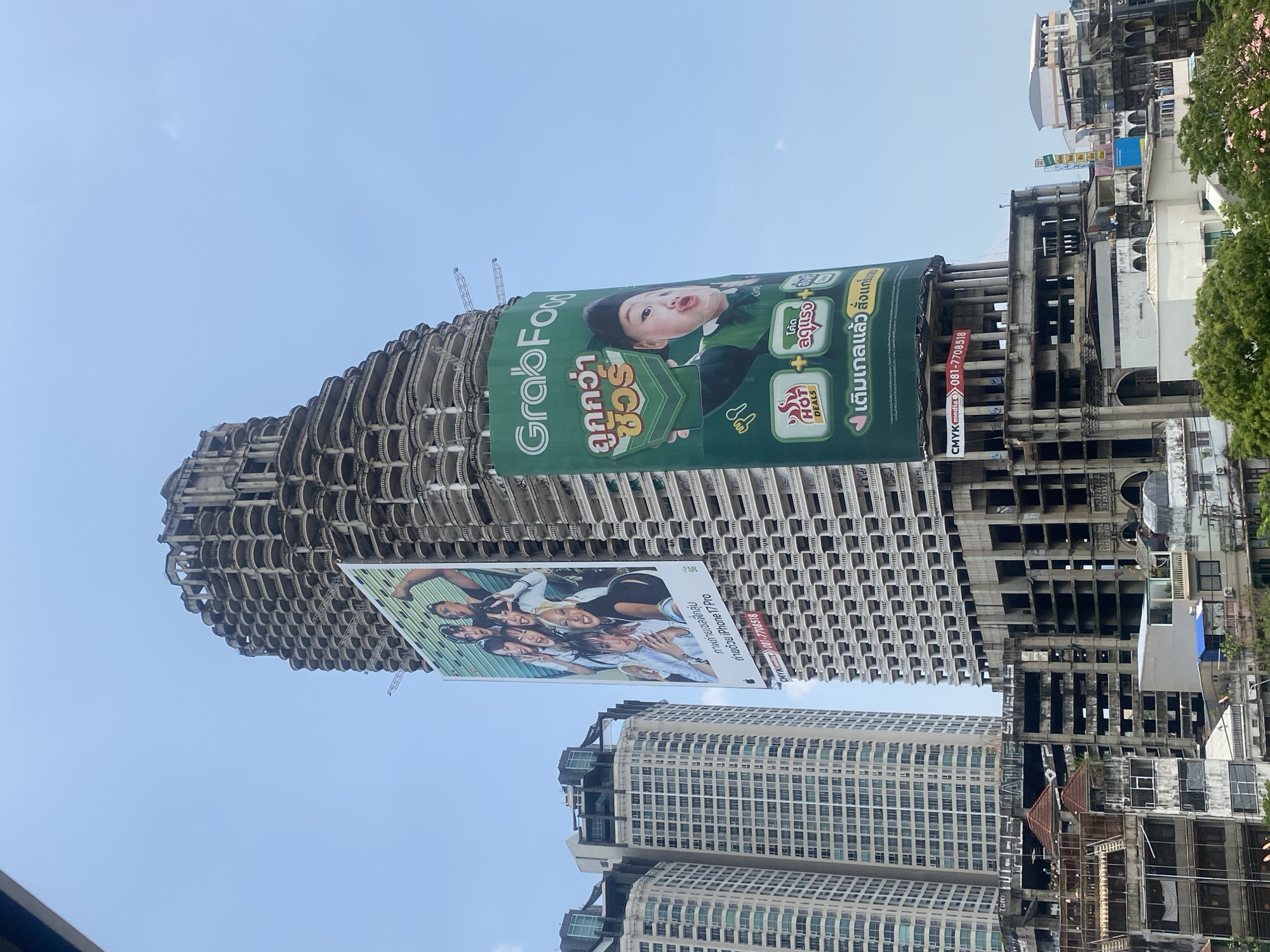 This is a medium shot of an unfinished skyscraper in Bangkok, Thailand, adorned with two large advertising billboards. The upper billboard is a green GrabFood advertisement featuring an infant holding a smartphone. The lower billboard displays an advertisement for a group selfie taken with an iPhone 17 Pro. The building appears to be in a state of disrepair with visible construction scaffolding. Other buildings are visible in the background, with a partially visible clear, light blue sky.
