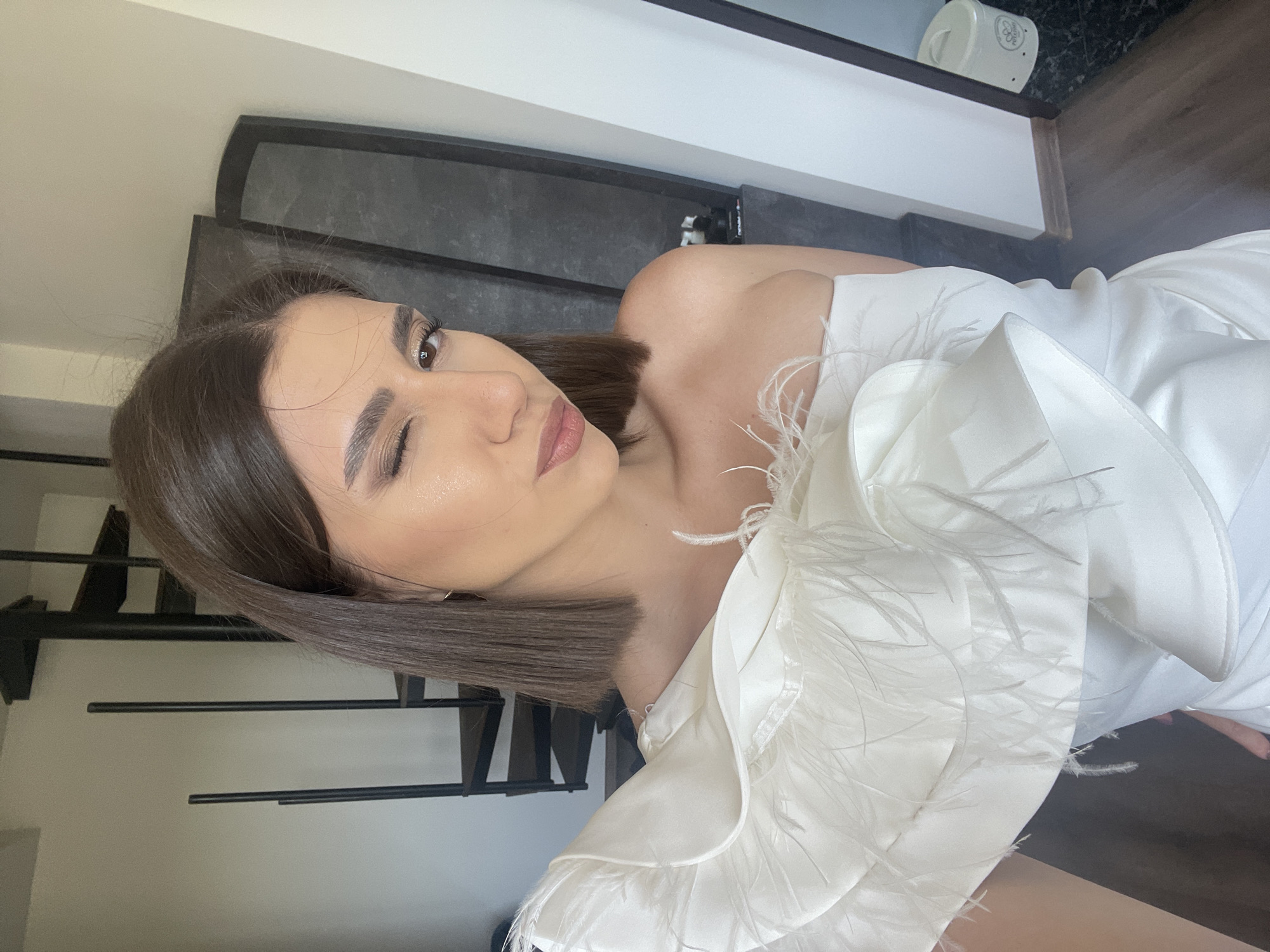 A woman with short brown hair, wearing a white dress with feather trim, is winking in the image. The background shows a white wall, a black and grey structure that might be a headboard, and a staircase with black metal railings. The location is likely indoors, but the city of Sofia, Bulgaria cannot be determined from the visual data.