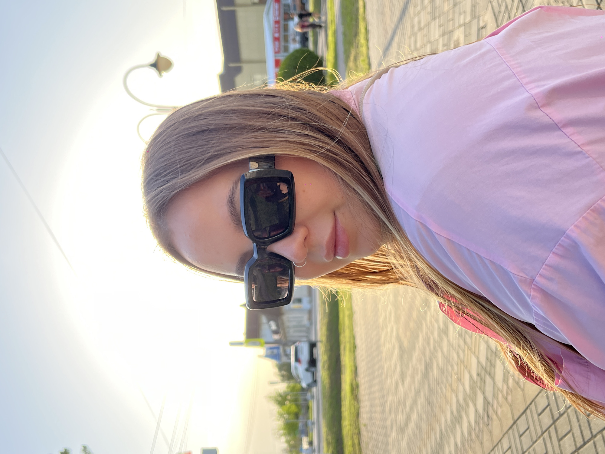 A young woman wearing sunglasses and a pink shirt is standing on a brick sidewalk. In the background, there are cars, trees, and buildings. The location is Novopokrovskaya, Russia.