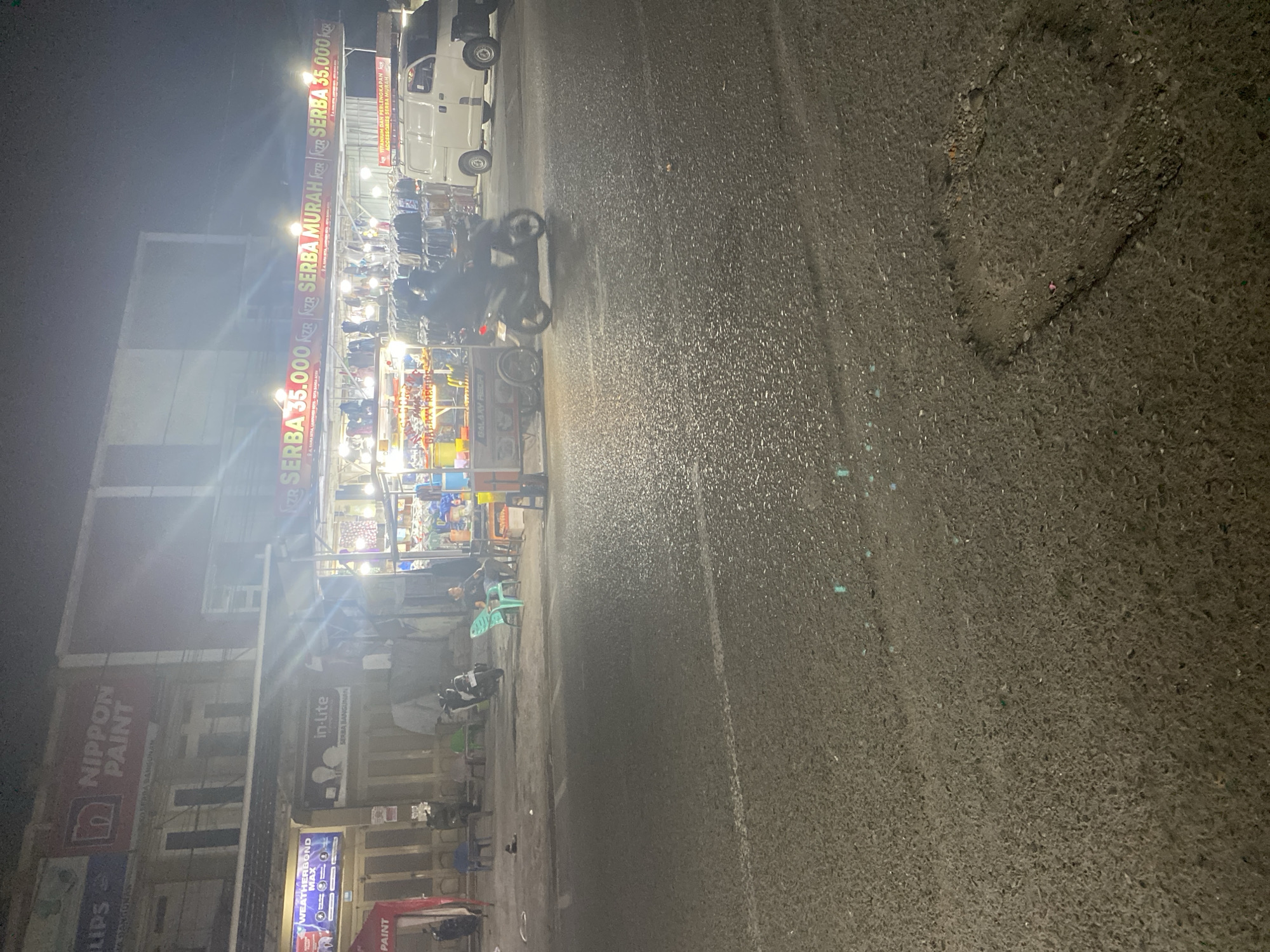The image depicts a nighttime street scene in Banda Aceh, Indonesia, likely near a commercial area. In the background, a building displays prominent signage for "NIPPON PAINT" and "in-lite SERBA BANGUNAN". Adjacent to this building, a row of stalls is illuminated, advertising "SERBA 35.000" (meaning "everything 35,000"). These stalls appear to be selling various items, possibly clothing and accessories, with racks of merchandise visible.
On the street in the foreground, a white van is parked to the right. A person is seated on a plastic chair on the sidewalk in front of the stalls. A motorcycle with two occupants is in motion on the street, heading towards the right of the frame. Another motorcycle is parked behind them. Several other people are visible seated or standing near the stalls. The road surface shows signs of wear and tear, with visible potholes. The lighting is a mix of artificial light from the stalls and streetlights, creating a somewhat hazy atmosphere.