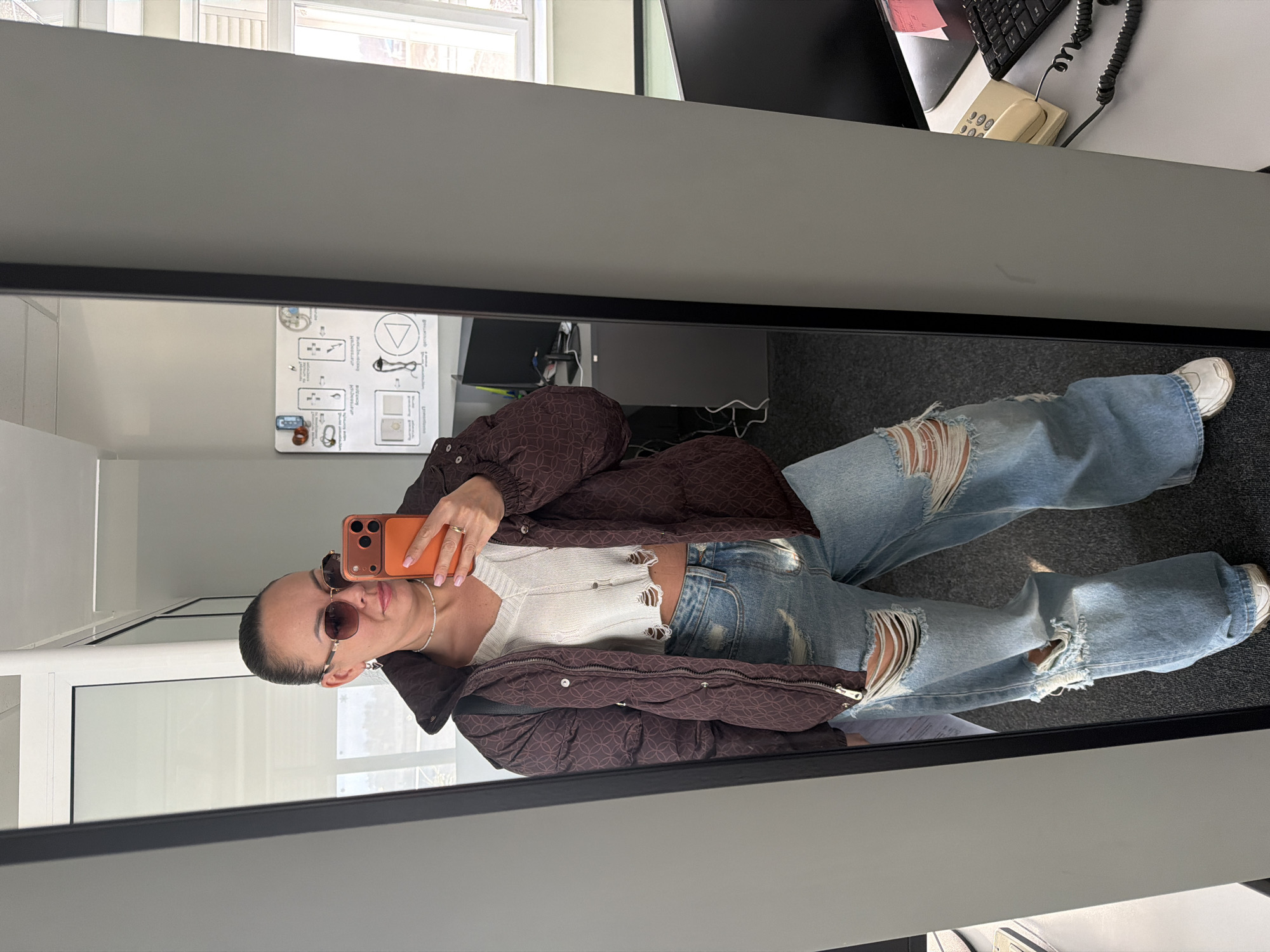 A woman stands in front of a full-length mirror taking a selfie. She is wearing sunglasses, a brown patterned puffer jacket, a white knit top, and ripped light blue jeans with white sneakers. She holds an orange cell phone in her right hand. The reflection reveals an office environment with a desk, computer, keyboard, and phone. There is a window with natural light. A diagram is visible on the wall. The location is unspecified and cannot be verified as Поляны, Russia from the visual data.