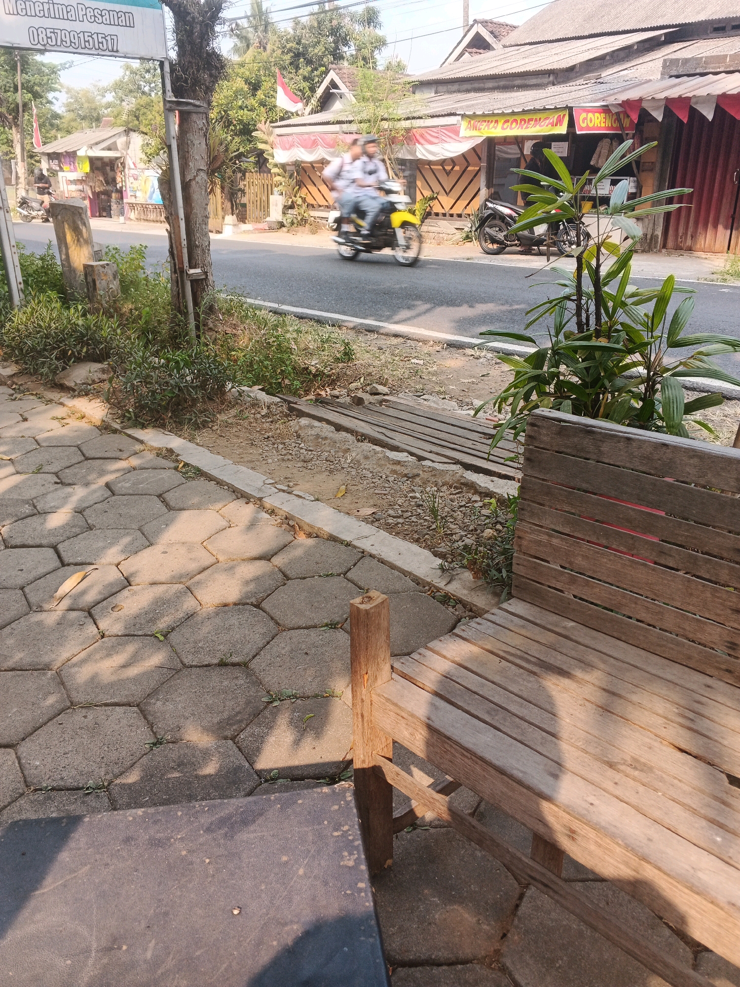 The image captures a street scene in Ungaran, Indonesia, likely in front of a food stall. A wooden bench is prominently positioned in the foreground, with a dark-colored surface visible in the lower left corner, possibly a table. The ground is paved with hexagonal concrete blocks. Beyond the bench, a road is visible with vehicles. A yellow motorcycle with two people on it is in motion, heading away from the viewer and towards a building with a corrugated metal roof. This building appears to be a shop or restaurant, with a banner that reads "ANEKA GORENGAN GORENGAN" (Various Fritters Fritters). Another motorcycle is parked in front of the building. Red and white flags are displayed, possibly indicating a national holiday or celebration. Green foliage is present throughout the scene, adding to the outdoor atmosphere. The lighting suggests it is daytime, with shadows cast by the objects.