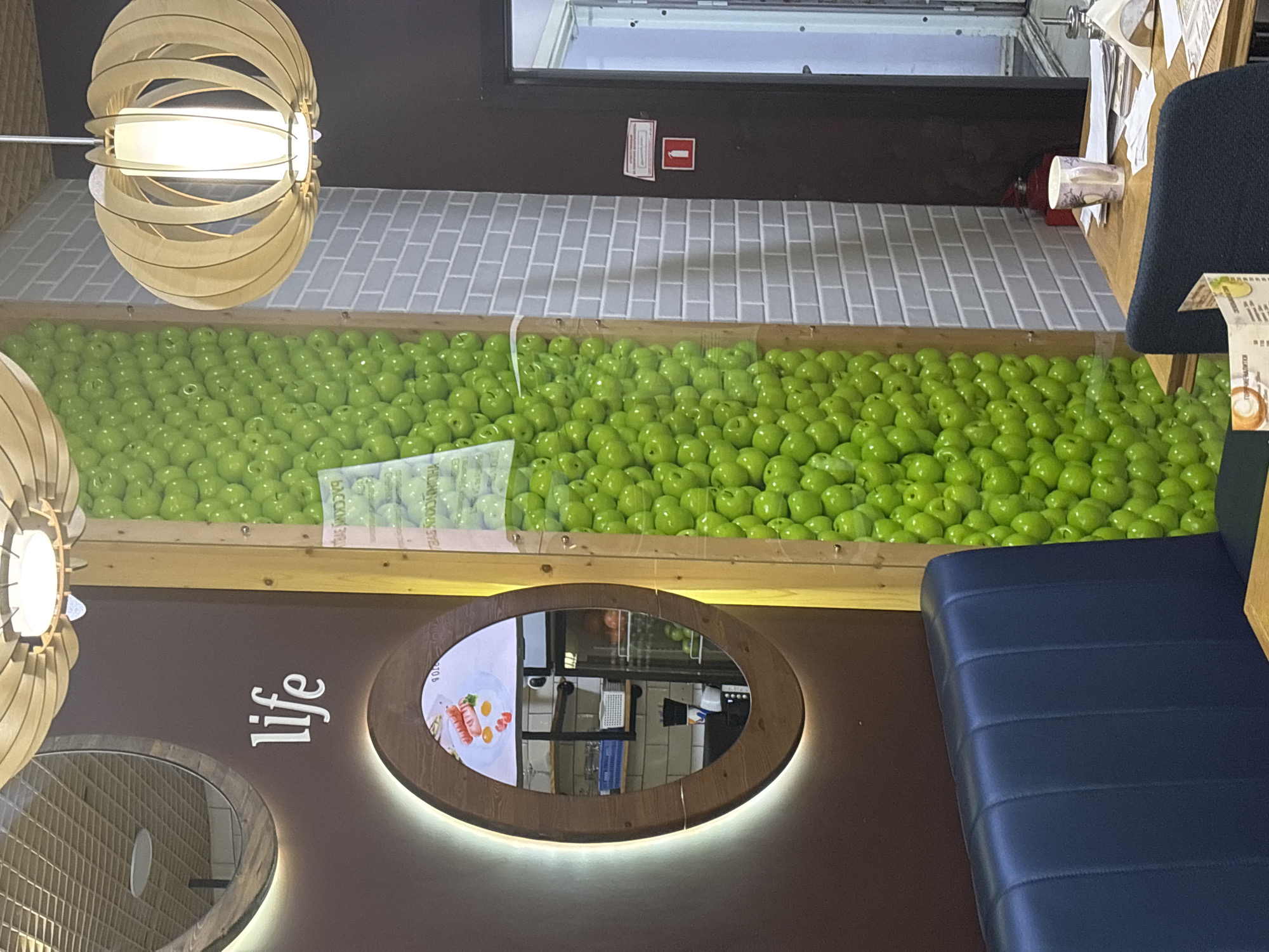 The image shows the interior of a cafe in Naberezhnye Chelny, Russia. A prominent feature is a wall decorated with a vertical arrangement of green apples. To the left, there is a dark brown wall with the word "life" spelled out in white lettering. Below this, an oval mirror with a wooden frame is illuminated by a soft glow. The mirror reflects a breakfast plate with sausages and eggs, as well as shelving units. A dark blue upholstered bench runs along the bottom left of the frame. To the right of the apple wall, a light grey brick wall is visible, and further right, a dark doorway leads to another area. A small red fire extinguisher is mounted on the brick wall, and a table with a mug and papers is partially visible in the foreground. The overall lighting is warm and inviting.