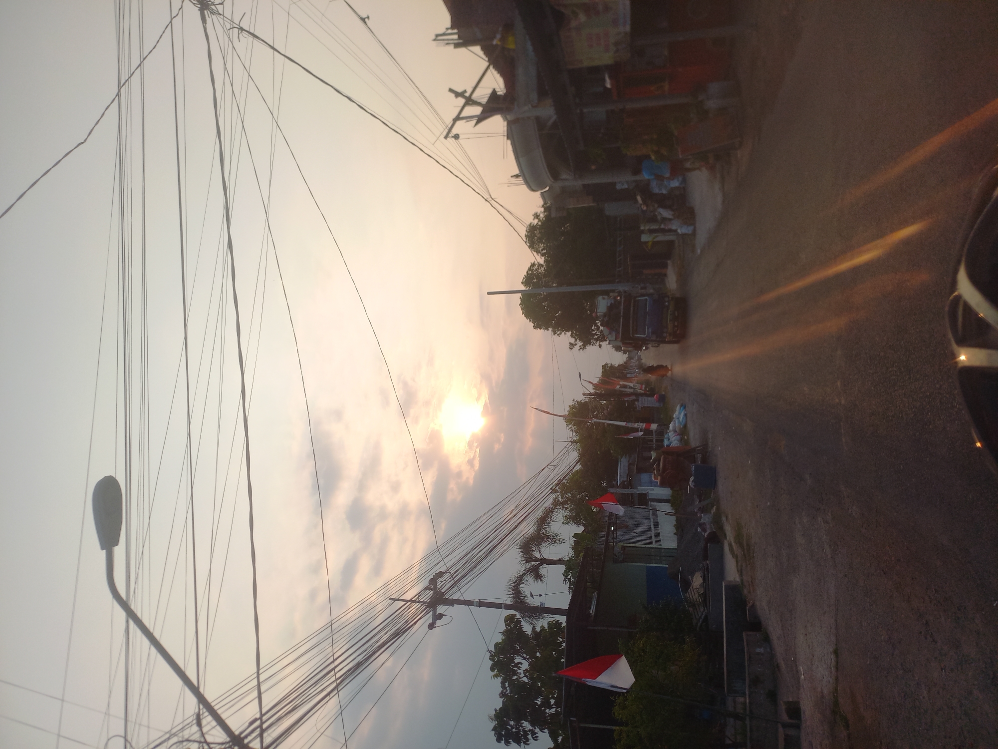 The image depicts a street scene in Bedah Lawak, Indonesia, during what appears to be late afternoon or early evening. The sky is overcast with the sun partially visible, casting a warm, diffused light and creating lens flare effects on the road. The street is unpaved and appears to be a residential or semi-commercial area.
On the left side of the street, there are several low-rise buildings, some with a corrugated metal roof. Indonesian flags are prominently displayed, indicating a local celebration or national holiday. Trees and vegetation are present, with some palm trees visible in the background. Utility poles with numerous electrical wires crisscross the scene, a common feature in Indonesian streetscapes.
On the right side of the street, more buildings are visible, some appearing to be shops or small businesses. Construction materials, such as sandbags and piles of gravel, are present, suggesting ongoing development or repairs. Several people are scattered throughout the scene, though their features are not clearly discernible due to the image quality and distance. A blue truck is driving down the street, and what appears to be a scooter or motorcycle is in the foreground, its handlebars visible. The overall impression is of a typical, somewhat busy, local street.