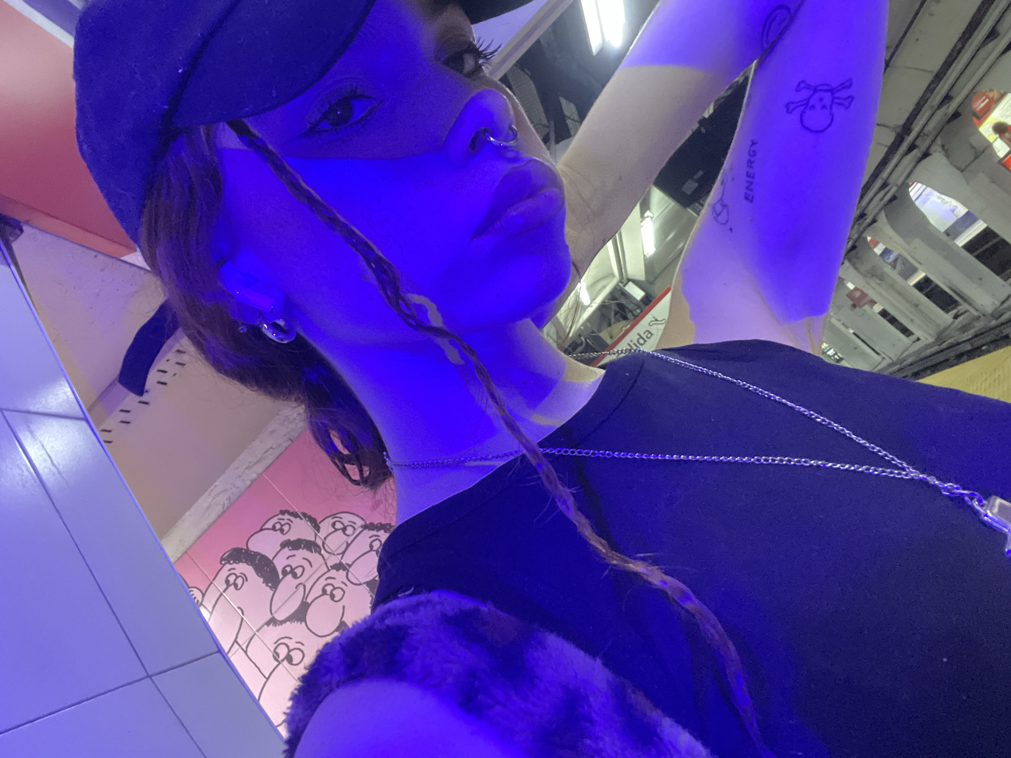 A close-up, low-angle shot taken under a blue light reveals a woman wearing a black baseball cap. She has a nose ring and an earring, and a single braid hangs down from her hair. She is wearing a black t-shirt with a silver necklace. The woman has tattoos on her arm. The background includes artwork with cartoonish figures on pink and white tiles, indicating she's likely in a subway station. A sign with "Salida" is visible. The location is likely the Buenos Aires subway system, known as Subte.
