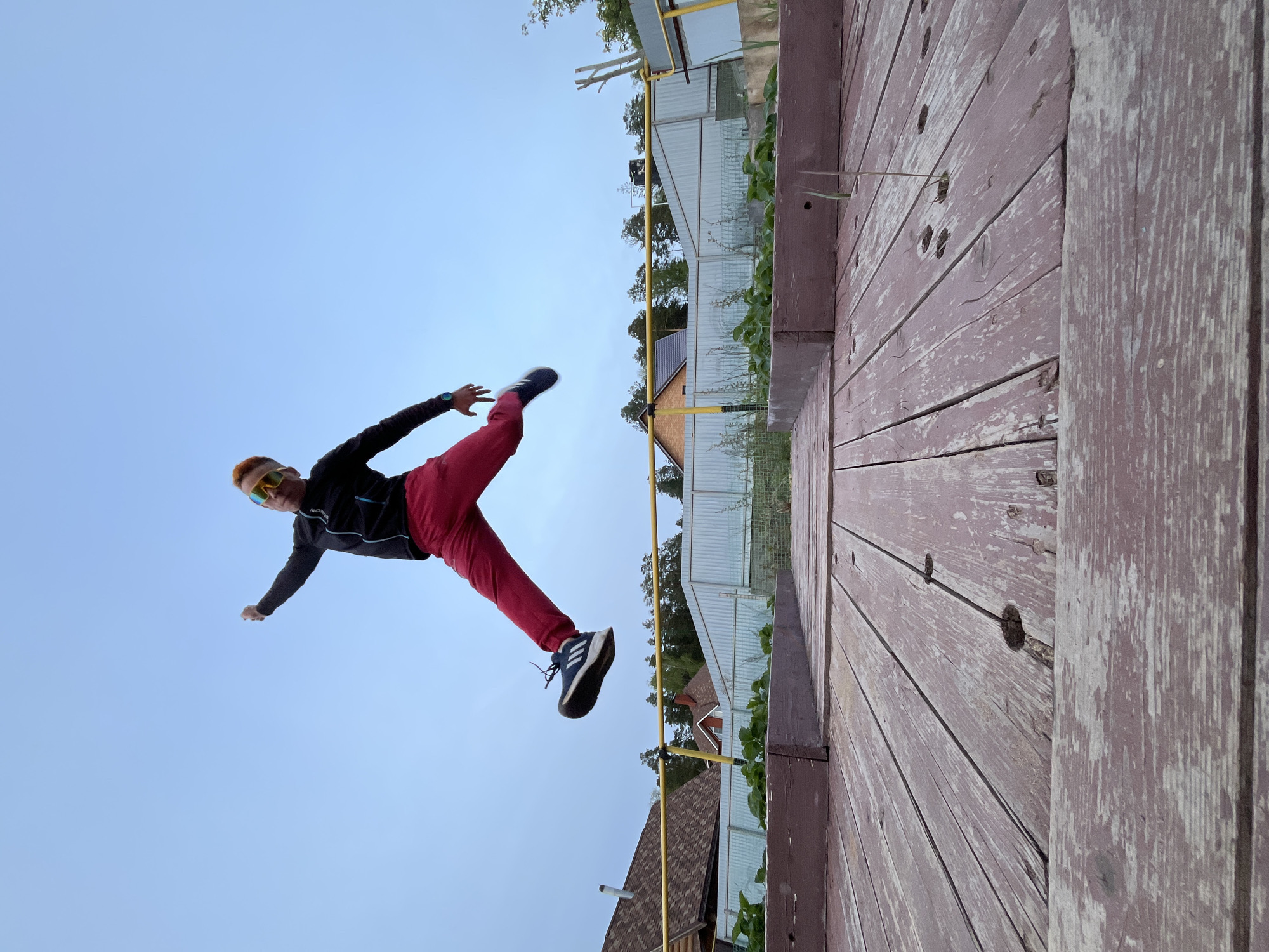 A person is captured mid-air, appearing to jump or kick, against a backdrop of a light blue sky. The individual is wearing red pants, a dark-colored athletic jacket, and athletic shoes, and sports orange hair and yellow-tinted sunglasses. The location seems to be Chaikovsky, Russia, as the architectural style of the houses and fences in the background is typical of this region. In the foreground, there's a wooden surface, possibly a deck or platform, with visible planks and weathered paint. The orientation of the image and the person's pose creates a dynamic and unconventional perspective.