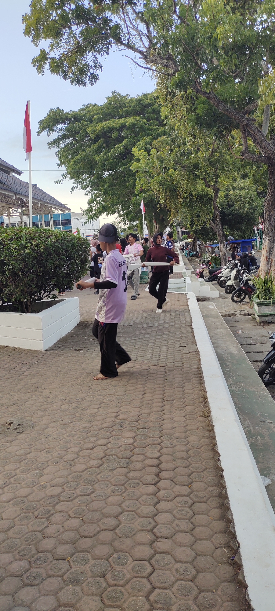 In Lhokseumawe, Indonesia, on a pedestrian walkway paved with hexagonal concrete blocks, a group of young people are engaged in an activity. The foreground features a young person wearing a light purple t-shirt with black sleeves and black pants, barefoot, and a dark grey baseball cap. This individual is holding a white baton. Behind them, other youths are also holding similar white batons, suggesting a synchronized movement or practice. Some individuals are wearing long-sleeved shirts in pink and maroon, along with dark pants. In the background, a building with a distinctive roof structure and a red and white flag are visible, indicative of a public space or institution. The environment includes mature trees with dense green foliage, a white-painted planter with a green shrub, and a row of parked motorcycles along the side of the walkway. The overall lighting suggests it is late afternoon or early morning, with a clear sky.