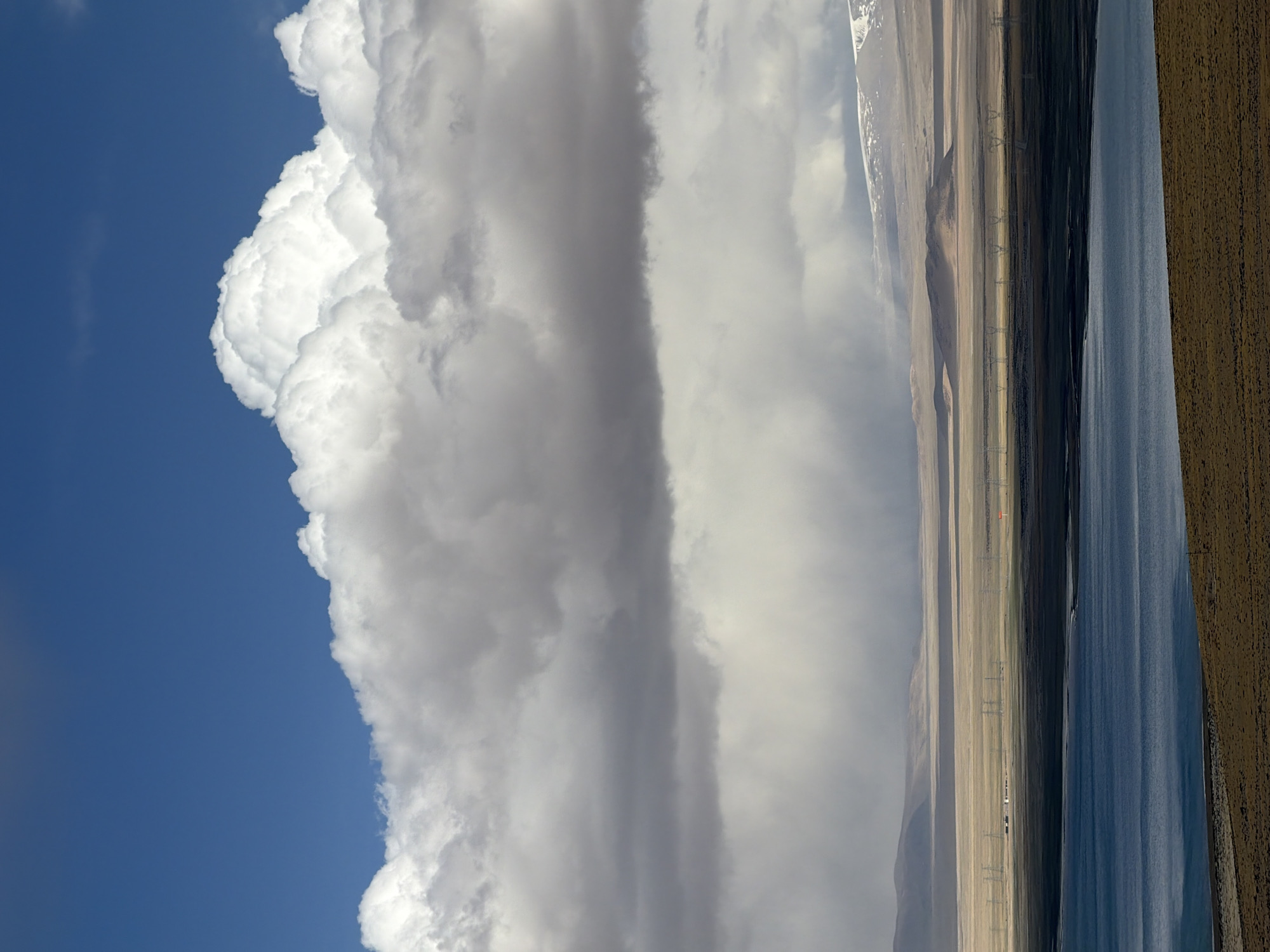 The image depicts a landscape in Gyirong County, China. A large cumulonimbus cloud dominates the left side of the frame, set against a blue sky. To the right of the cloud formation, a column of grey clouds extends towards the horizon. The right side of the image features a vista of mountains, arid plains dotted with power lines, a dark lake, and brown vegetation in the foreground. The image is oriented vertically, with the sky on the left and the ground on the right.