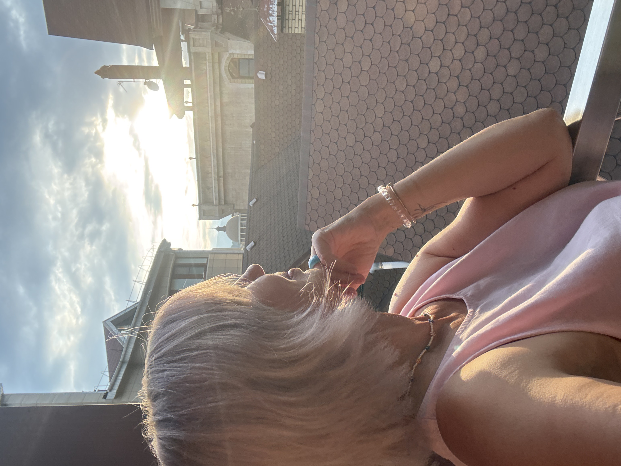 A woman with blonde hair, wearing a pink tank top and a beaded necklace, is seen in profile, looking out at a city skyline at sunset. Her left hand is resting on her chin, and she is wearing bracelets on her left wrist. The background shows rooftops, buildings, and a cloudy sky with the sun breaking through. The location is specified as Estosadok, Russia.
