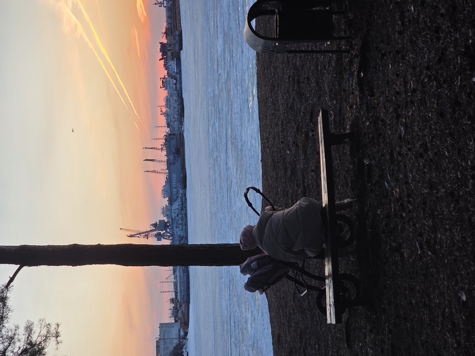 A low-angle shot of a person sitting on a park bench next to a stroller near the Kama River in Chaikovsky, Russia, facing a series of boats and industrial buildings. The scene is framed by a tree on the left and a trash can on the right. The river appears to be partially frozen. The sky is painted with hues of orange and yellow, suggesting either a sunset or sunrise. Airplanes are leaving contrails in the sky.