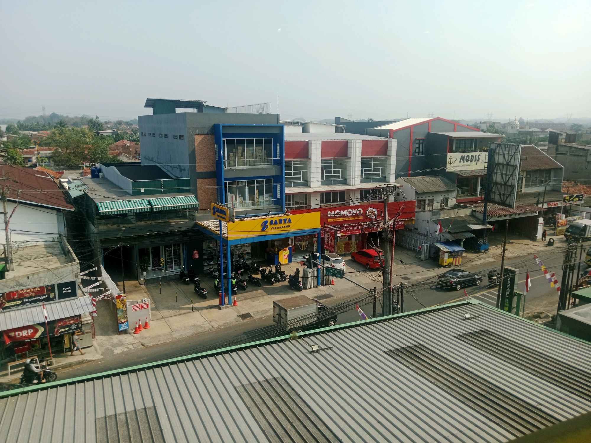 The image shows a street scene in Ciranjang, Indonesia, on a clear day. In the foreground, a corrugated metal roof is visible. The middle ground features a row of buildings along a street. A prominent yellow and blue building with the text "SATYA CIRANJANG" is visible, appearing to be a store. Next to it is a building with red and white accents and the text "MOMOYO". Several motorcycles are parked in front of these establishments, and a few people are seen walking. Further down the street, a red car and a white car are parked, along with a black car driving away. A white truck with a cargo box is also on the street. In the background, more buildings and trees are visible, indicating an urban or suburban environment. The sky is hazy.