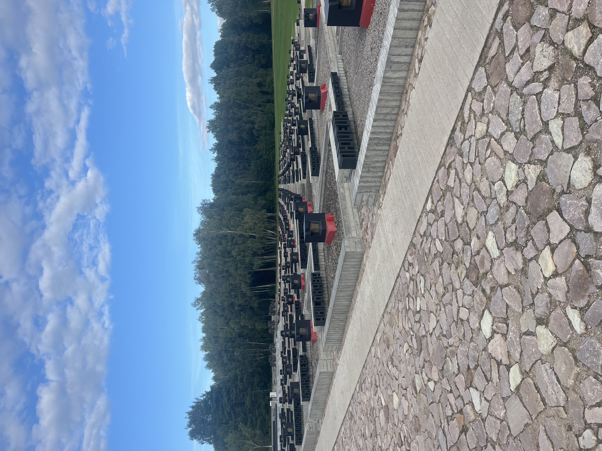 The image depicts a memorial cemetery with rows of identical grave markers. The markers are black and rectangular, some with red accents at the top. They are arranged in a grid pattern on a gravel base, bordered by concrete structures. A paved pathway runs through the cemetery. In the background, a dense line of trees forms a horizon, and beyond that, a blue sky with scattered white clouds is visible. The overall impression is one of solemnity and remembrance. This appears to be the memorial complex at the Khatyn memorial, located in the Logoisk District of Minsk Region, Belarus, rather than in the city of Каменскі сельскі Савет, Belarus.