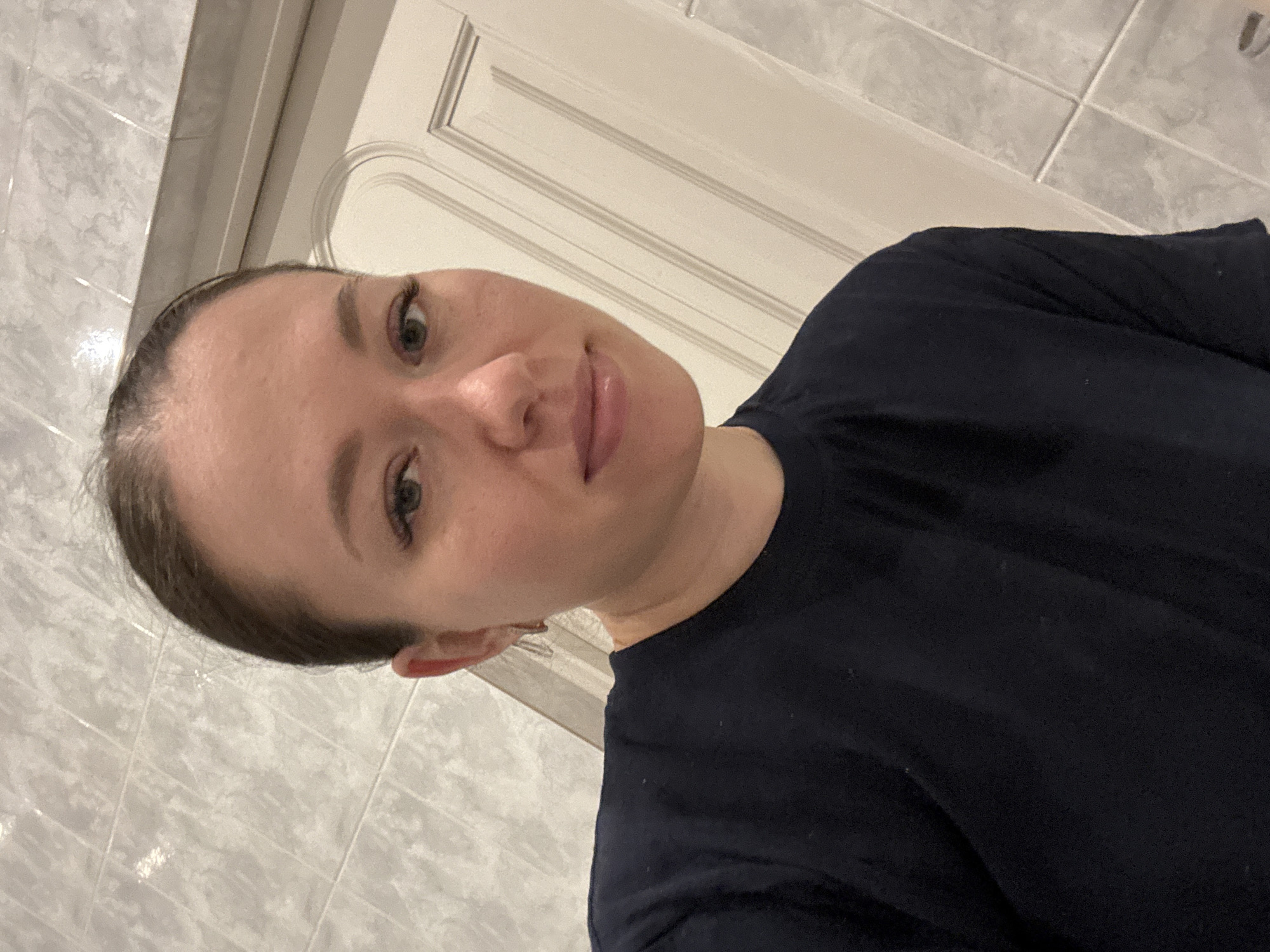 A woman with fair skin and dark hair pulled back is looking directly at the camera. She is wearing a dark blue or black t-shirt. She has light-colored eyes and her lips appear to have a slight gloss.
The background consists of tiled walls and a paneled door or wall with white trim. The location is inferred to be Ферма, Russia.