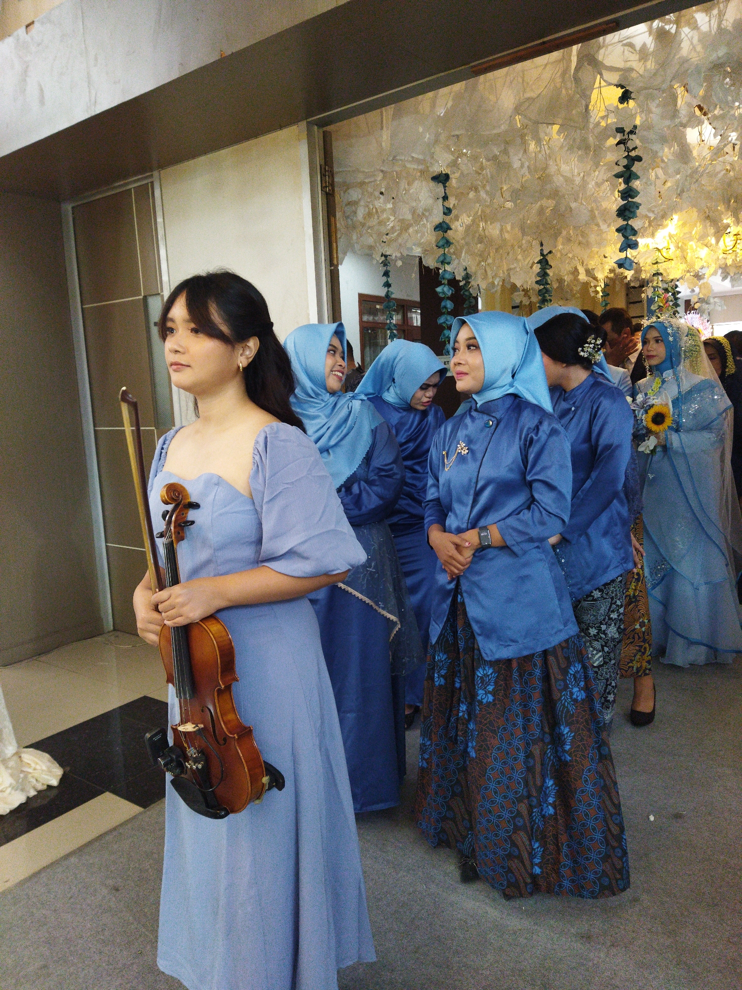 In Semarang, Indonesia, a young woman, possibly a wedding guest or performer, stands in the foreground holding a violin and bow. She is wearing a light blue, floor-length dress with puffed sleeves and a sweetheart neckline. Behind her, a group of women, also in blue Islamic attire, are lined up, some wearing hijabs. The woman second from the front is wearing a blue top and a patterned skirt with blue and brown floral motifs. Further back, a bride in a pale blue wedding dress and veil is visible, holding a sunflower bouquet. The background features a decorative archway with white floral arrangements and hanging blue accents, suggesting an indoor event like a wedding reception or ceremony. The flooring appears to be carpeted. The lighting is warm and appears to be artificial.