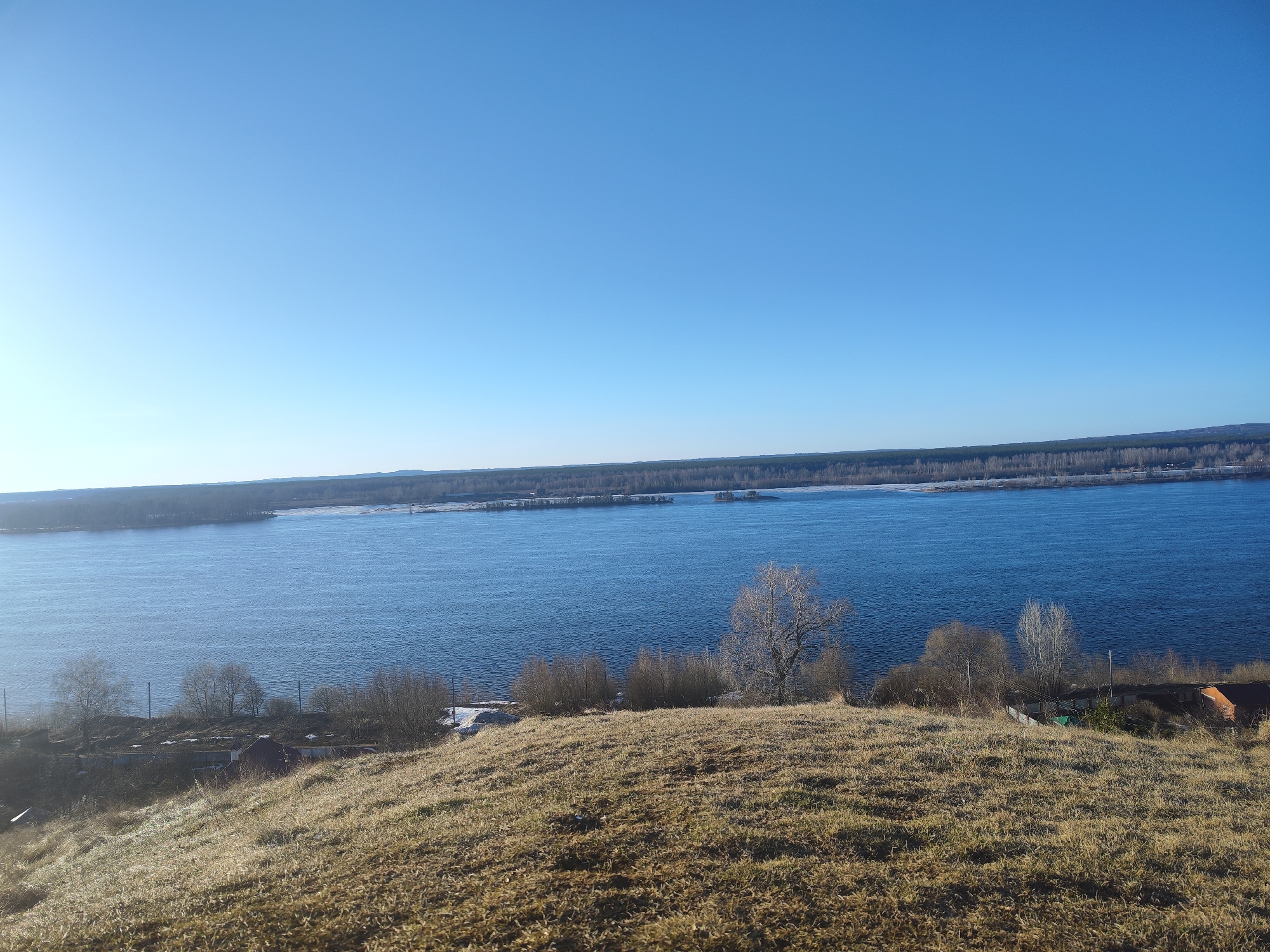 The image depicts a wide river landscape under a clear blue sky. The river, likely the Volga, occupies the middle ground, with its surface reflecting the sky. A line of trees borders the opposite bank. In the foreground, a grassy hillside slopes down toward the river. Sparse vegetation and bare trees are visible on the hillside. Some buildings can be discerned among the trees on the far right. The scene is bathed in sunlight, suggesting a daytime setting. The photograph was taken in Noviy, Russia.