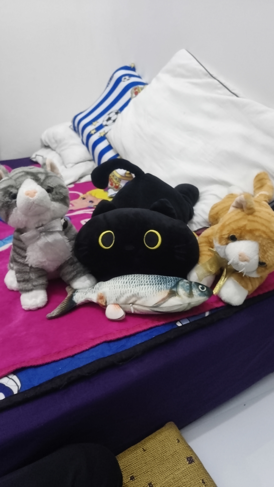 The image displays a collection of plush toys, including three cats and a fish, arranged on a bed. A gray and white striped cat is on the left, a black cat with large yellow eyes is in the center, and an orange tabby cat with a golden ribbon is on the right. The fish plush toy is positioned in front of the black cat. The bed is covered with a pink blanket and a blue and white striped pillow with soccer ball motifs is visible in the background. The scene is set indoors, likely in a bedroom. There are no people present in the image. The overall lighting suggests an indoor environment with some natural light. The image does not contain any identifiable landmarks or features that would indicate a specific location within Betung, Indonesia.