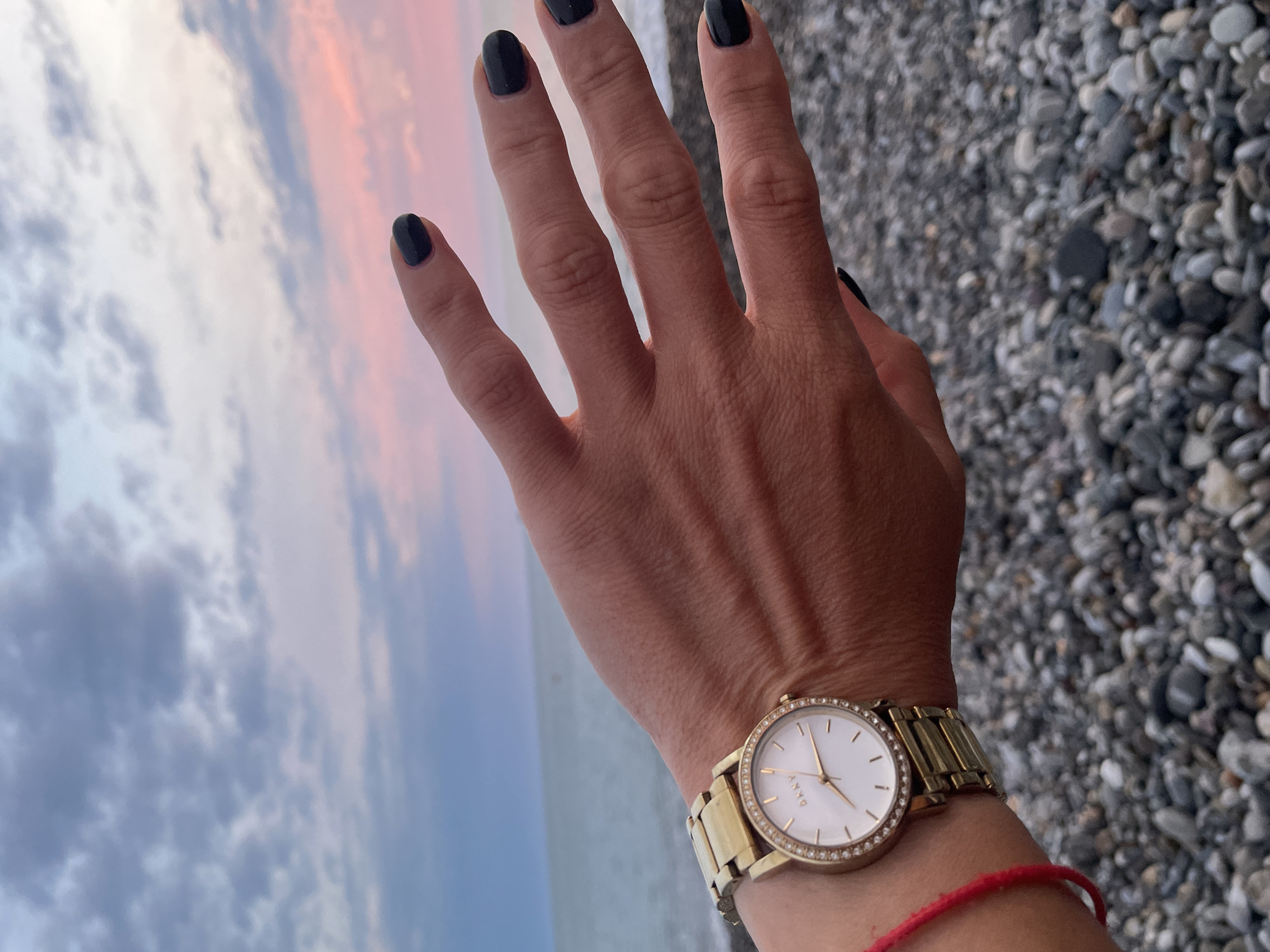 A close-up shot focuses on a woman's left hand and wrist against a backdrop of a pebble beach, the sea, and a dusky sky. The hand is extended, with fingers slightly spread, and its nails are painted in a dark, glossy polish. A gold-colored watch with a white face and diamond-like accents on the bezel adorns the wrist, with a thin red string bracelet visible beneath it. The sea stretches out to the horizon, meeting a sky filled with soft hues of pink, purple, and blue clouds, indicative of twilight or dawn. The ground in the foreground is covered with a dense scattering of smooth, grey and white pebbles.